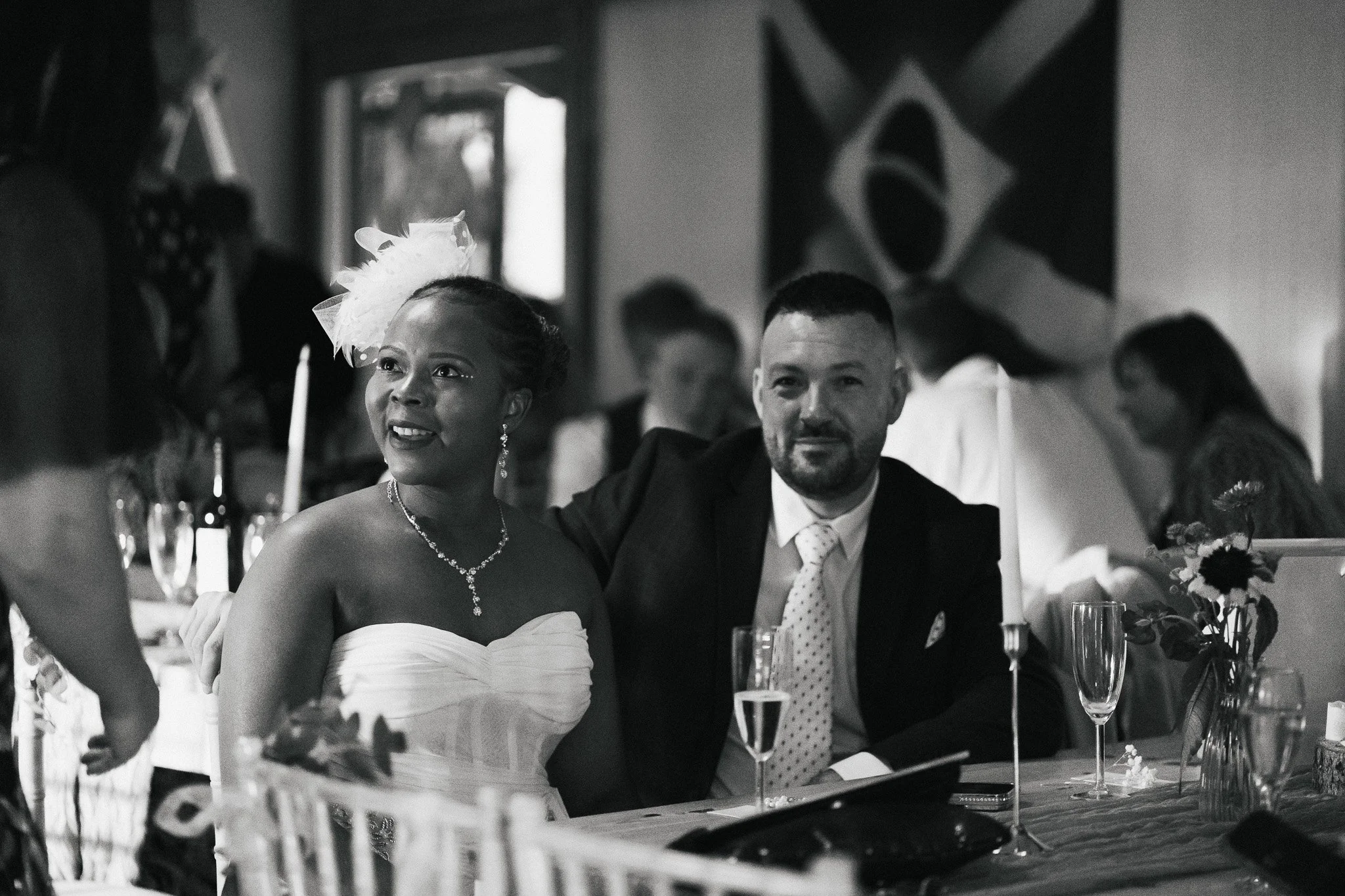 A woman in a wedding dress and a man in a suit sitting together at a wedding reception. The woman is wearing a necklace and earrings, with her hair styled up and a fascinator in her hair. The man has a beard and mustache, and is wearing a tie with po