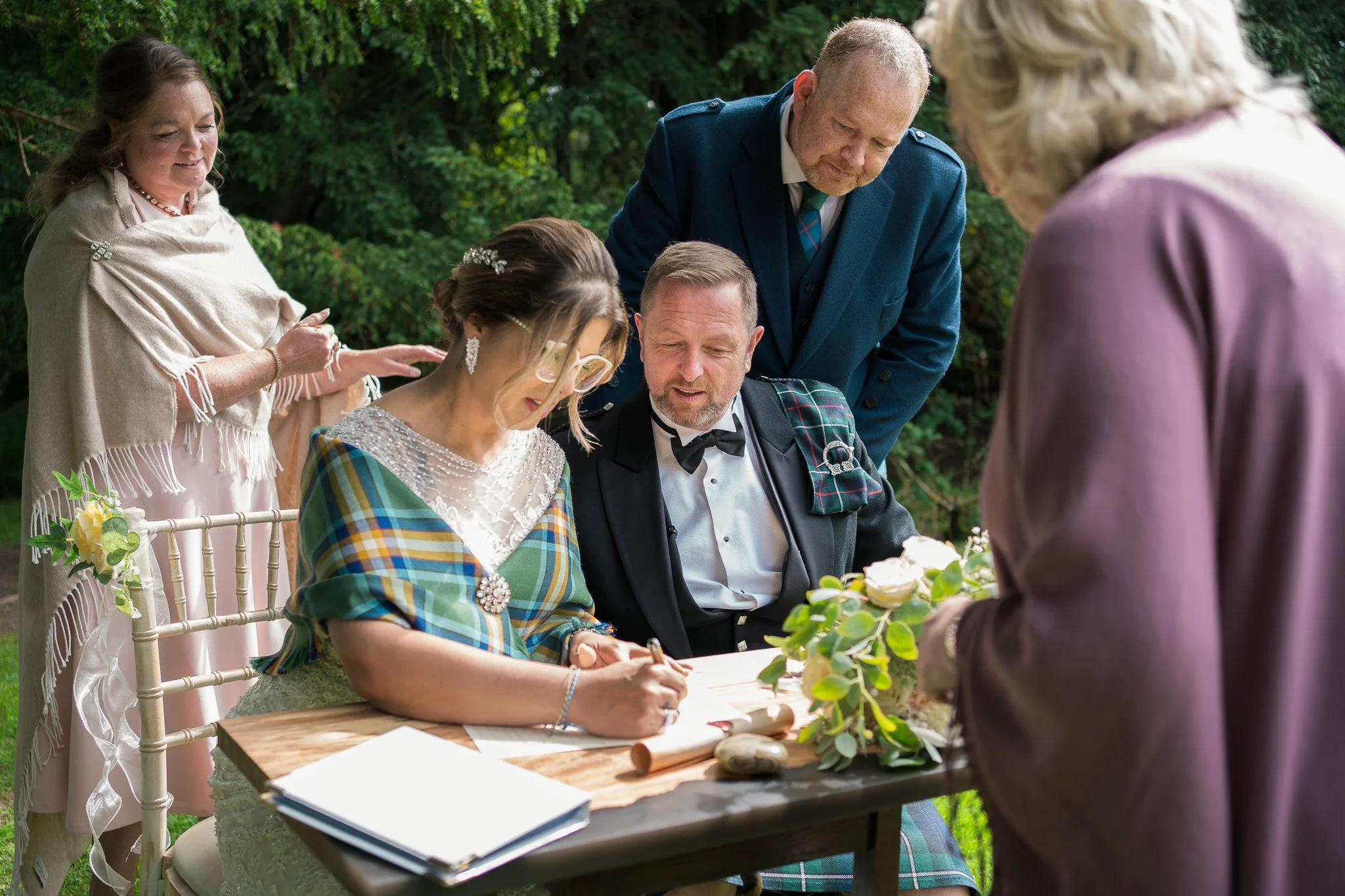 A wedding ceremony taking place outdoors with the bride signing a document at a wooden table, surrounded by family and friends.