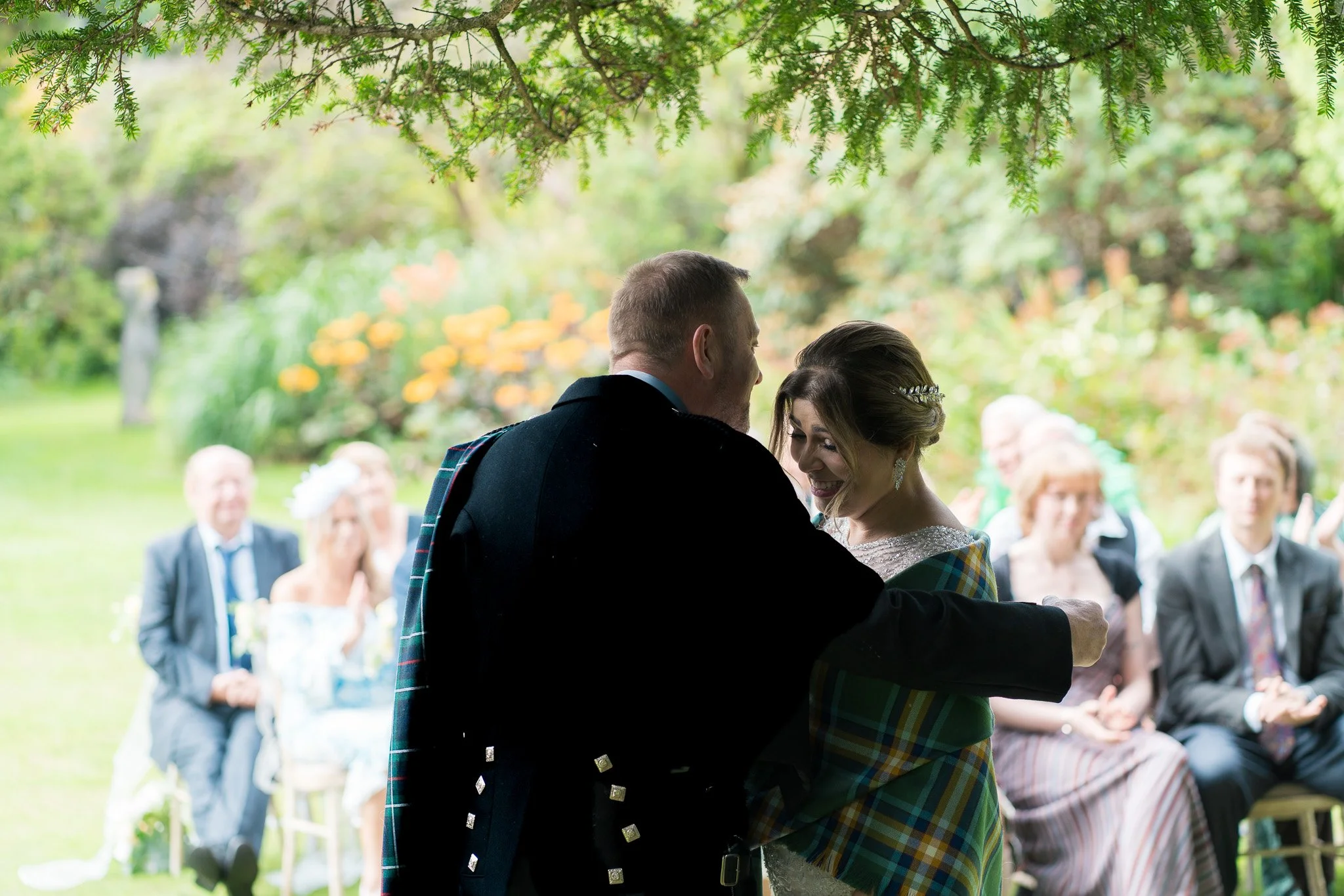 A bride and groom sharing a kiss during their outdoor wedding ceremony, with guests seated in the background under a leafy tree.