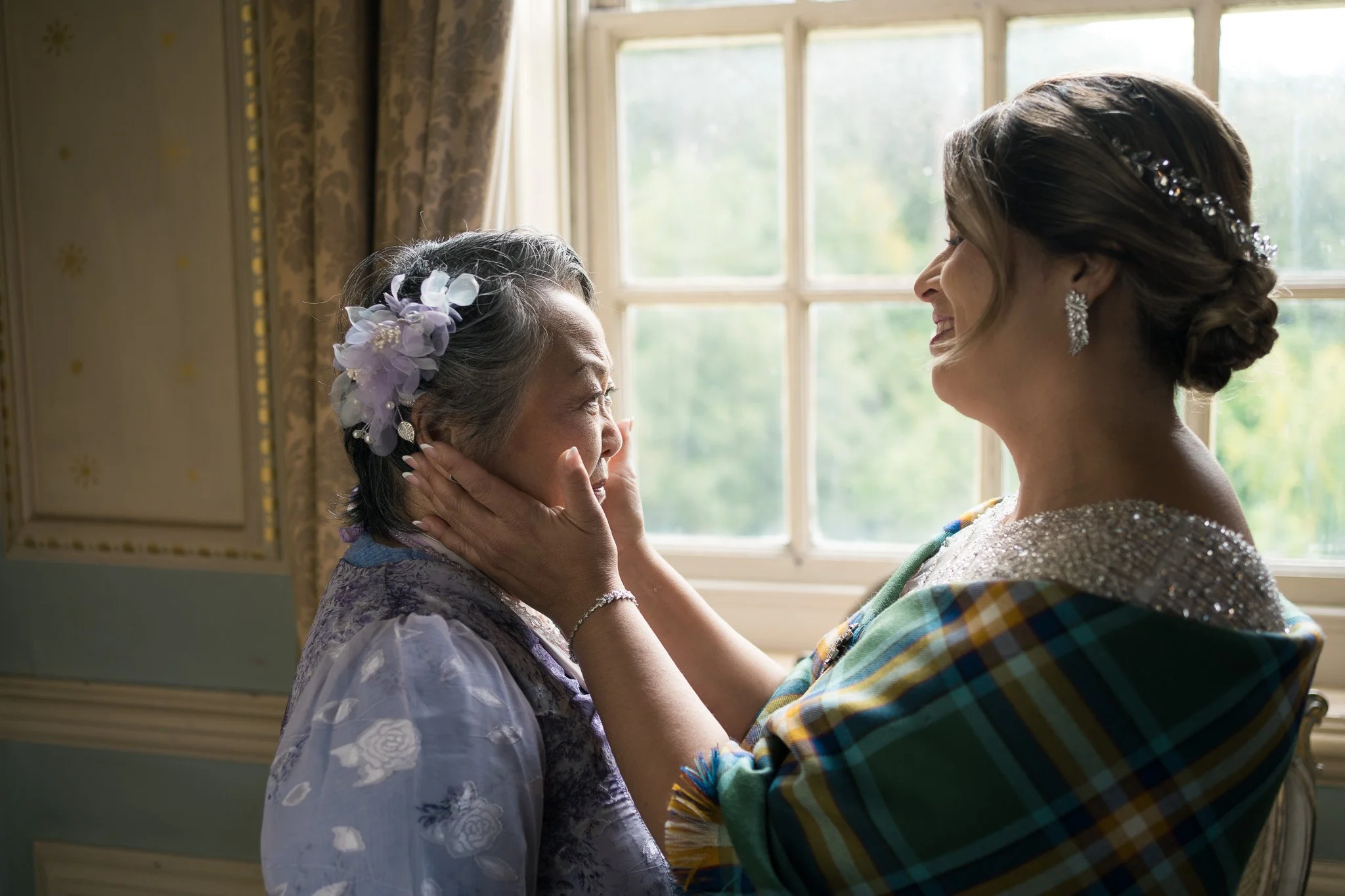 A woman and an elderly woman are sharing a tender moment by a window, smiling at each other. The elderly woman wears a lavender flower hair accessory, and the woman wears elegant earrings and has an ornate hairstyle with a decorative hair clip. The l
