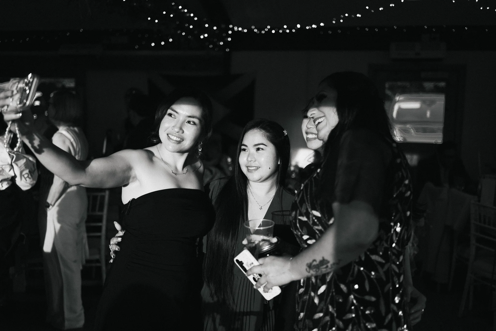 Four women are taking a selfie together at a social event. One woman holds a smartphone to capture the photo, and they are smiling and looking at the camera. The photo is in black and white.