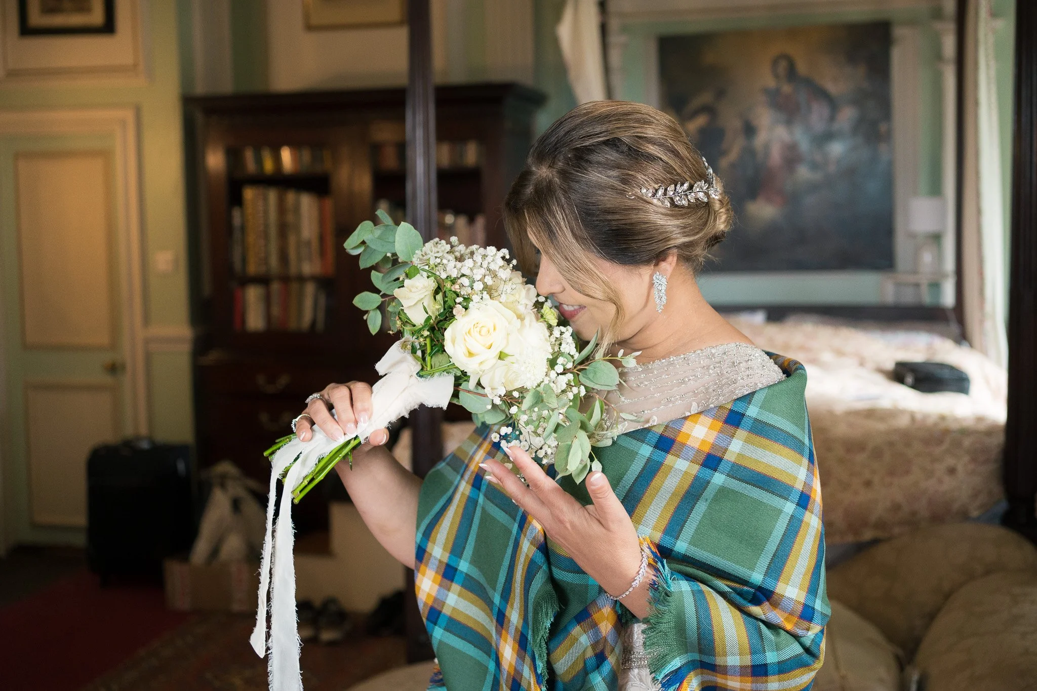 A woman dressed in a wedding gown with pearl earrings and a decorative hairpiece holds a bouquet of white roses, baby's breath, eucalyptus, and greenery, standing indoors near a bookcase and artwork.