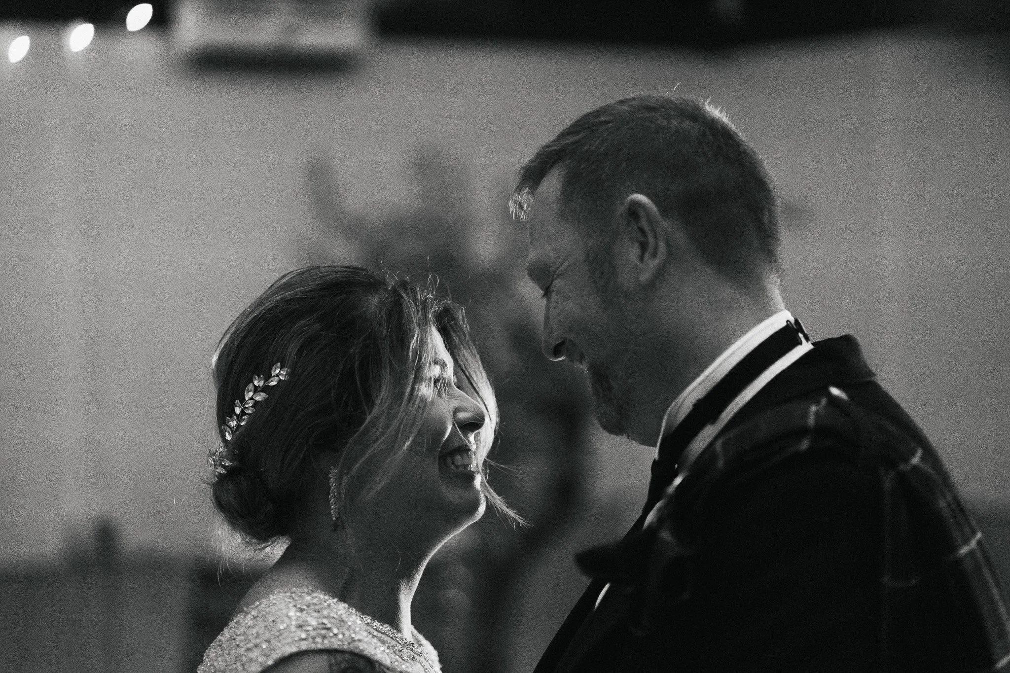 A black and white photo of a smiling woman and a man facing each other closely, possibly at a formal event, with blurred background lights.