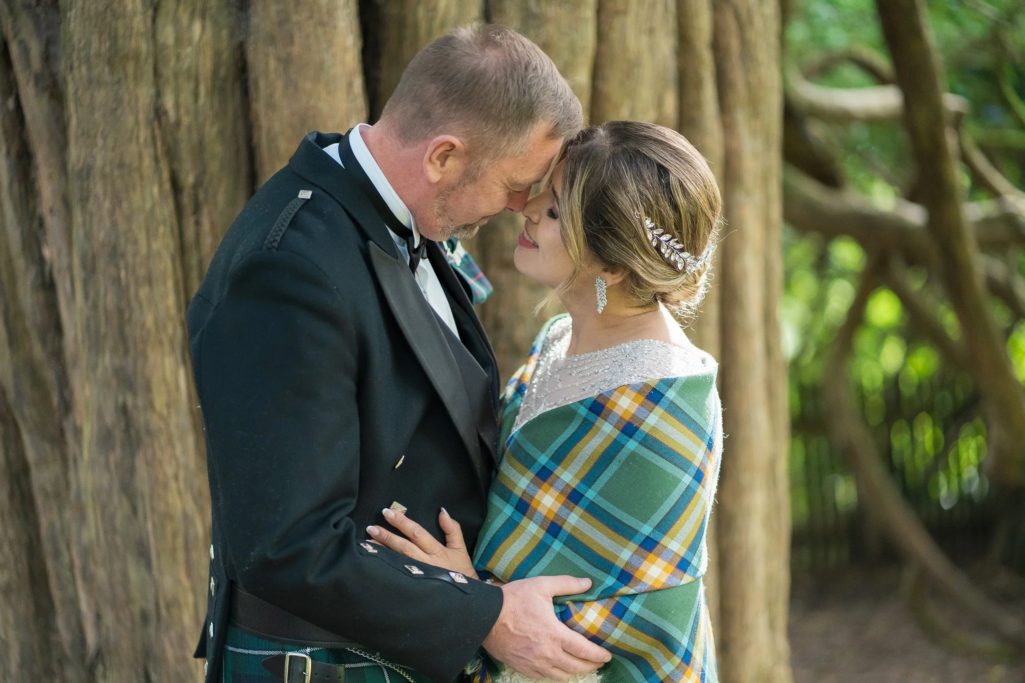A couple from different cultural backgrounds dressed in wedding attire, holding hands and touching foreheads, standing outdoors with a wooden fence and green trees in the background.