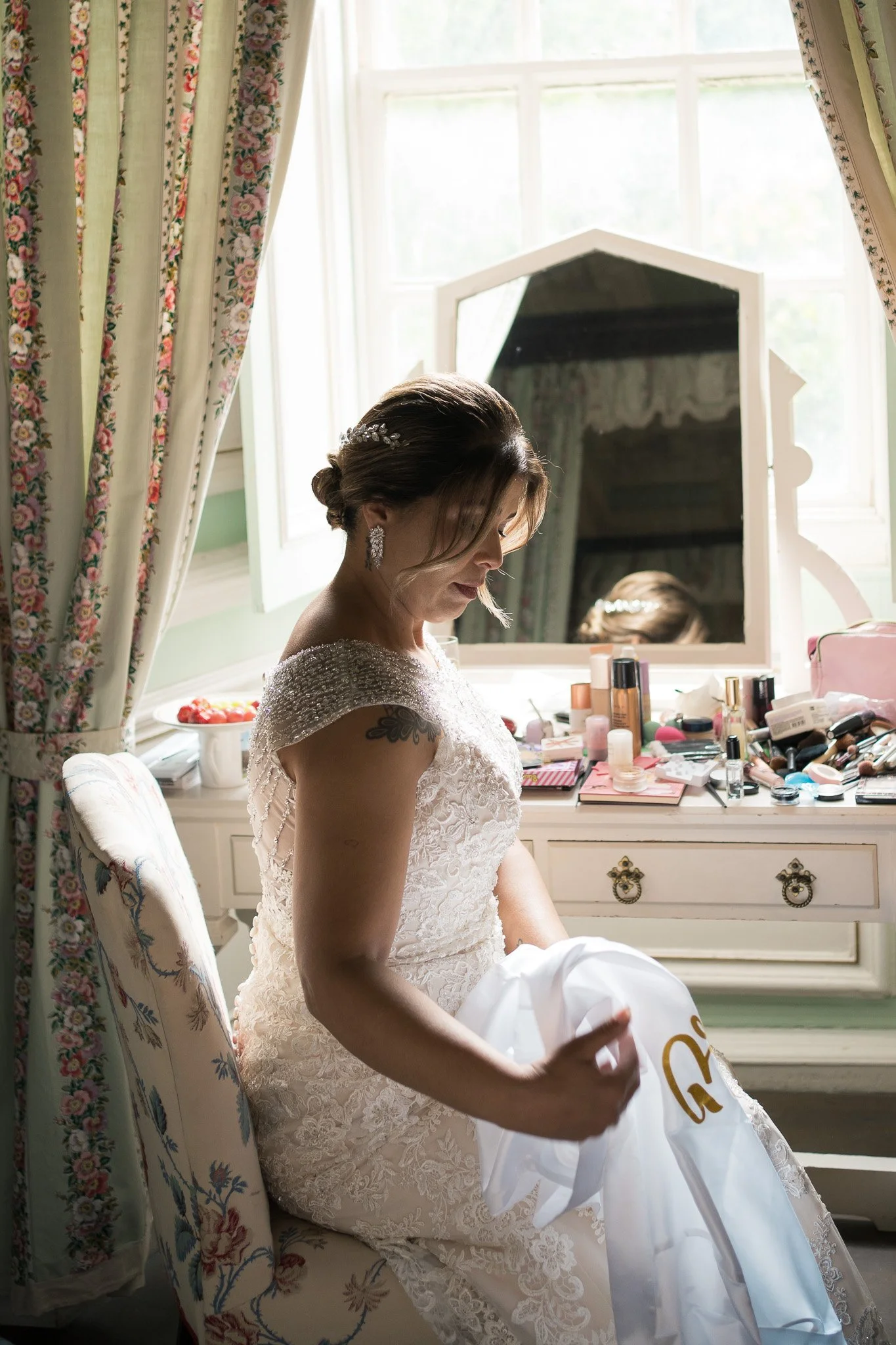 A woman dressed in a wedding gown sitting on a floral armchair, in front of a dresser with various makeup items. She is holding a white bag with gold lettering, with a mirror and window with floral curtains in the background.