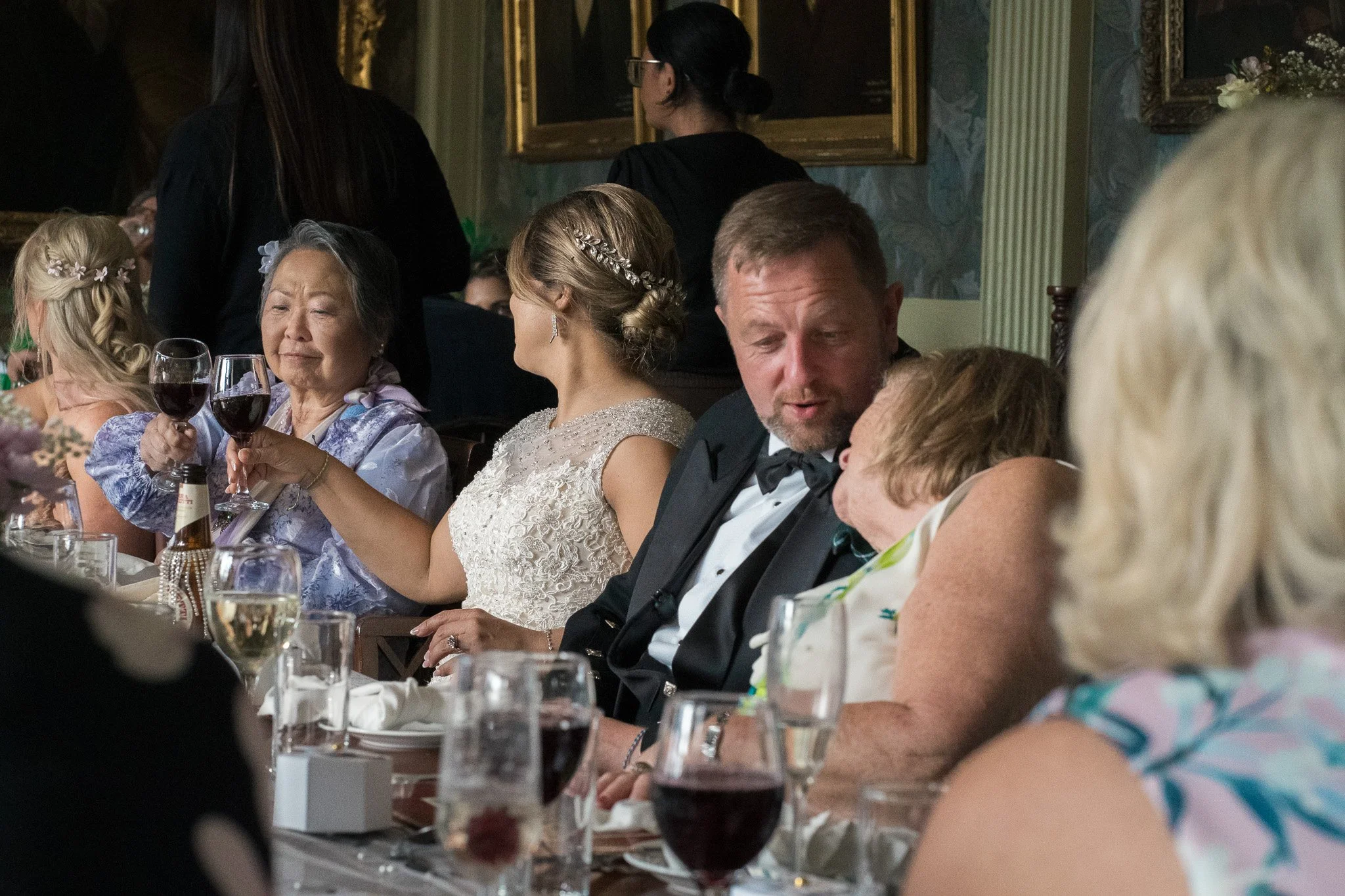 People sitting at a formal dinner table, including an elderly woman holding a glass of red wine, a woman in a white dress, and a man in a tuxedo, with paintings on the wall behind them.