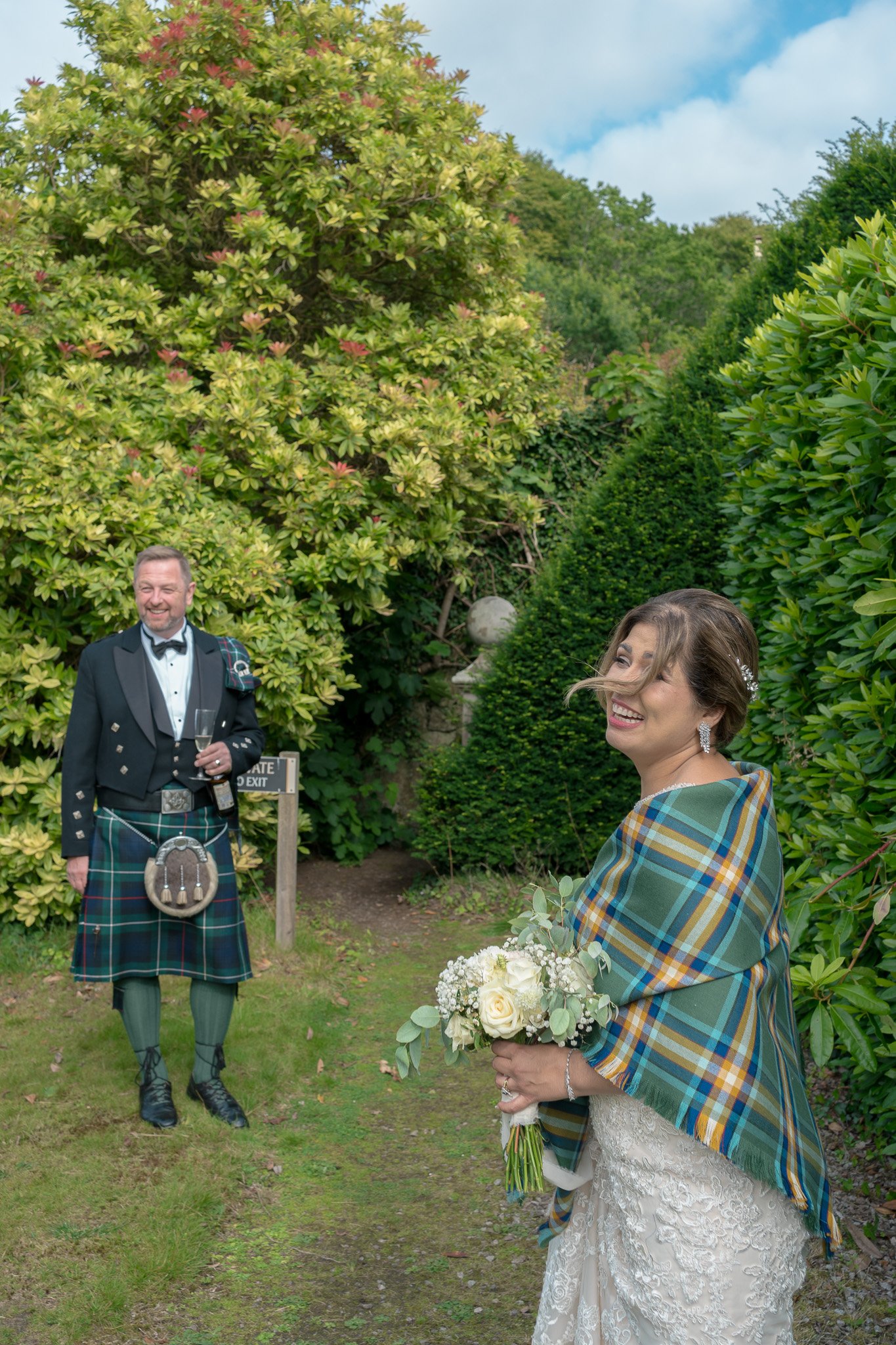 A bride holding a bouquet of white flowers and a woman draped in a tartan blanket inside a garden with lush greenery, including large bushes and trees, and a stone pathway. A man in traditional Scottish attire with a kilt and sporran stands in the ba
