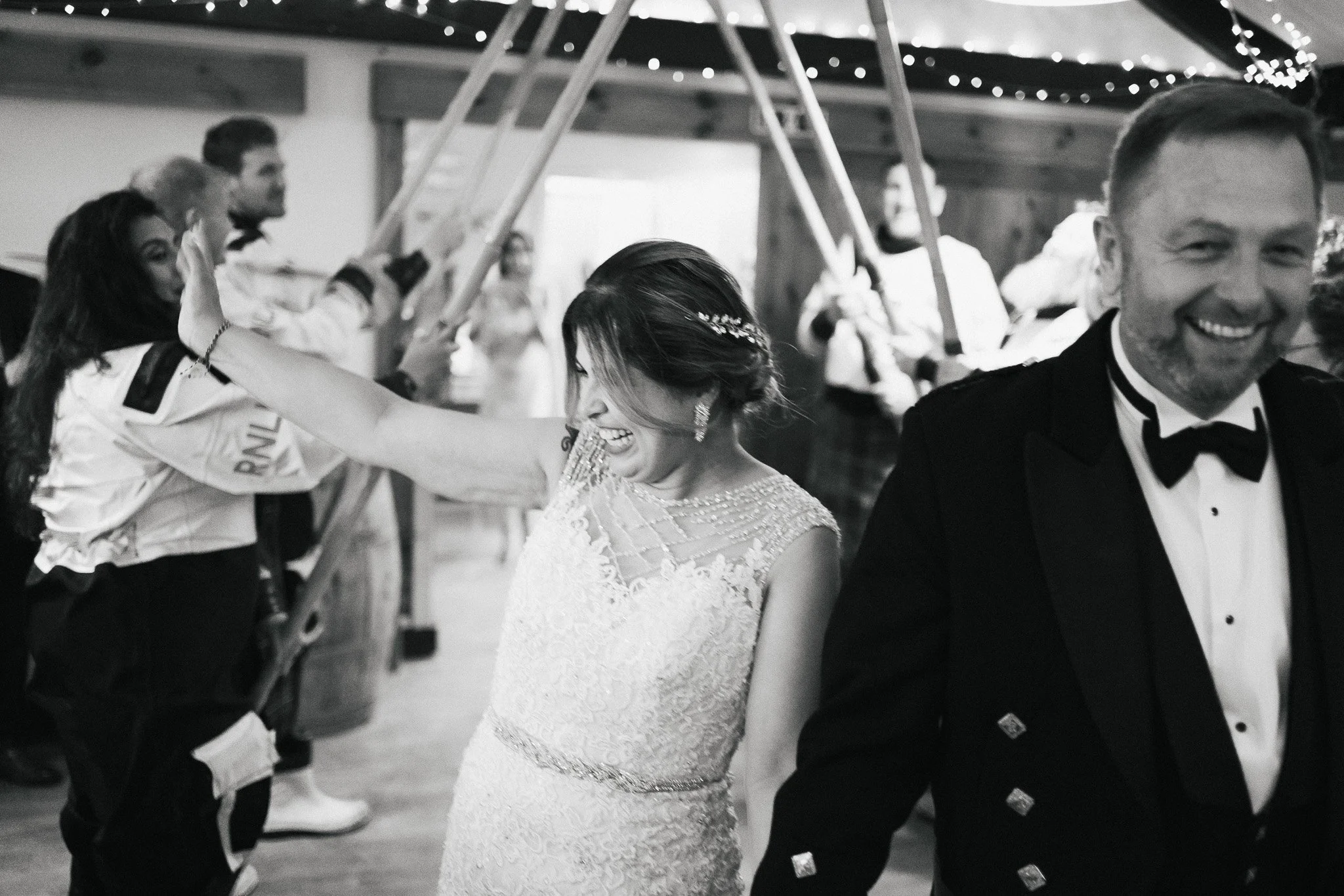 A black and white photo of a wedding reception where a bride and groom are dancing. The bride is smiling and wearing an elegant lace wedding dress with a beaded hair accessory. The groom is dressed in a tuxedo and is also smiling. In the background, 