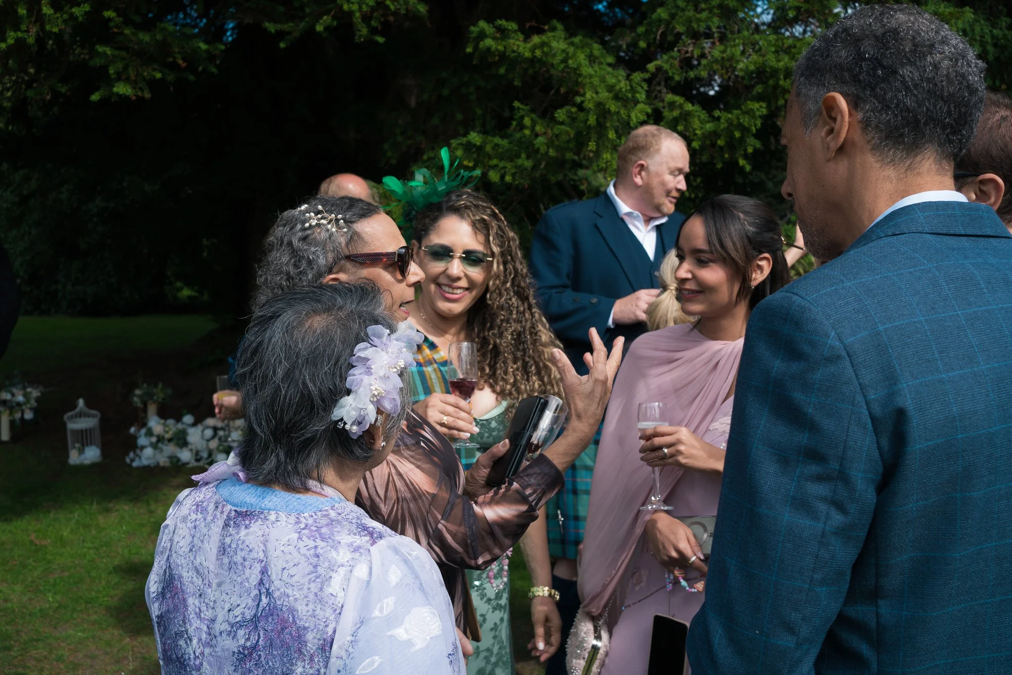 A group of people at an outdoor celebration, dressed in elegant attire, with some holding drinks, talking, and smiling in a garden setting with greenery and decorative flowers.