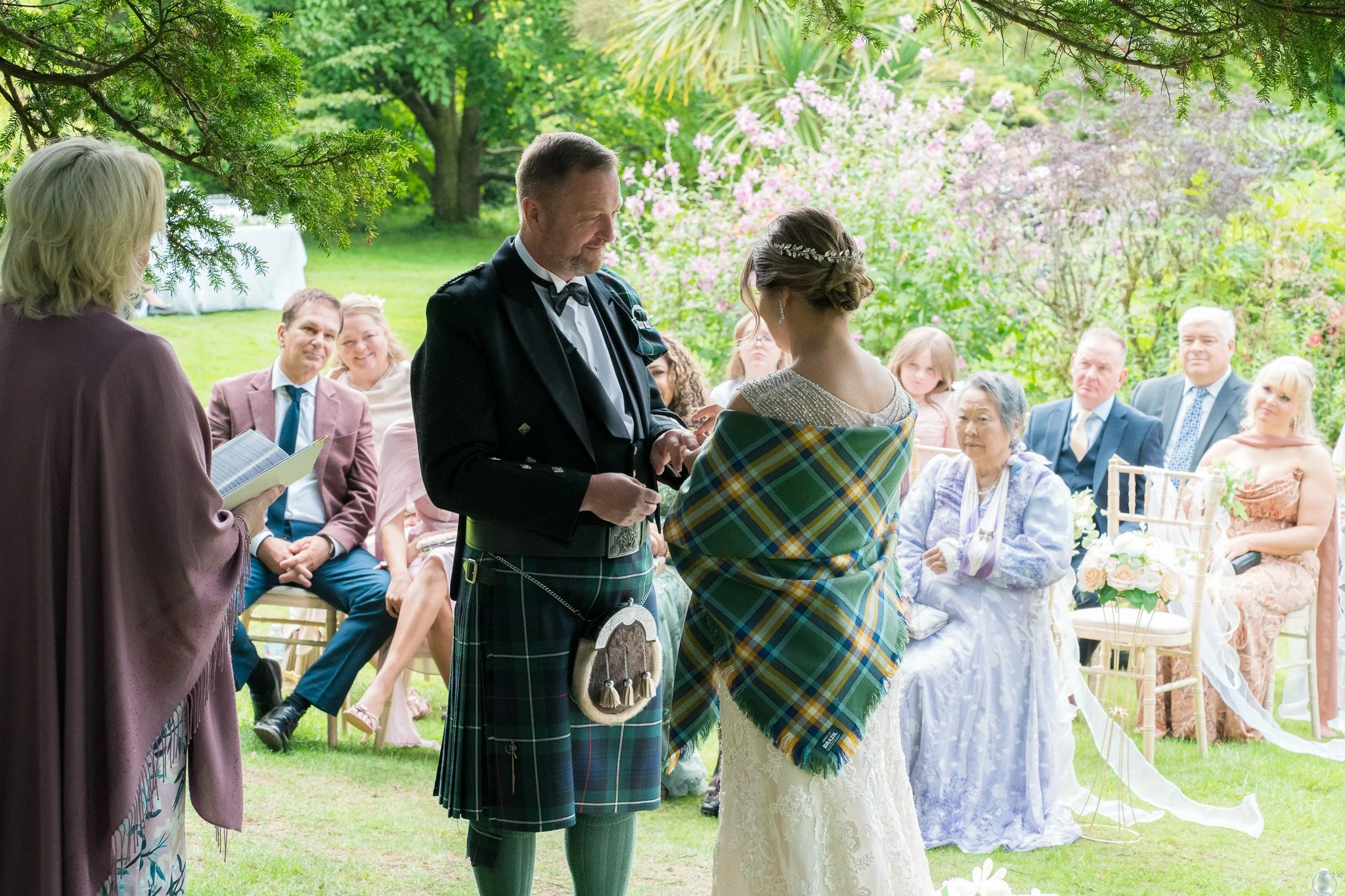 A wedding ceremony outdoors with a groom in traditional Scottish kilt and a bride in a white dress, surrounded by seated guests under a green canopy with pink flowering trees in the background.