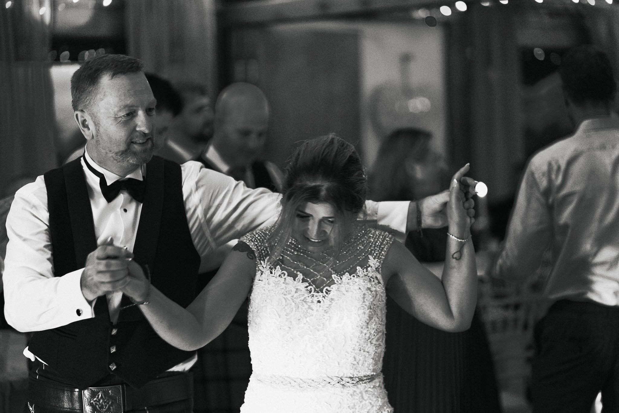 A black and white photo of a man and woman dancing at a formal event. The man is wearing a tuxedo with a bow tie and has his arm around the woman's shoulder. The woman is wearing a lace dress and is smiling while holding onto the man. There are other