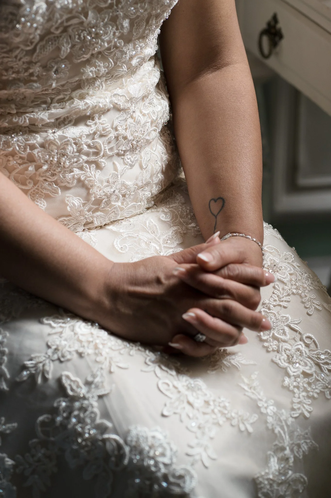 Close-up of a bride in a wedding dress with lace and embroidery, showing her left arm with a small heart tattoo and a silver bracelet, while her hands are clasped.