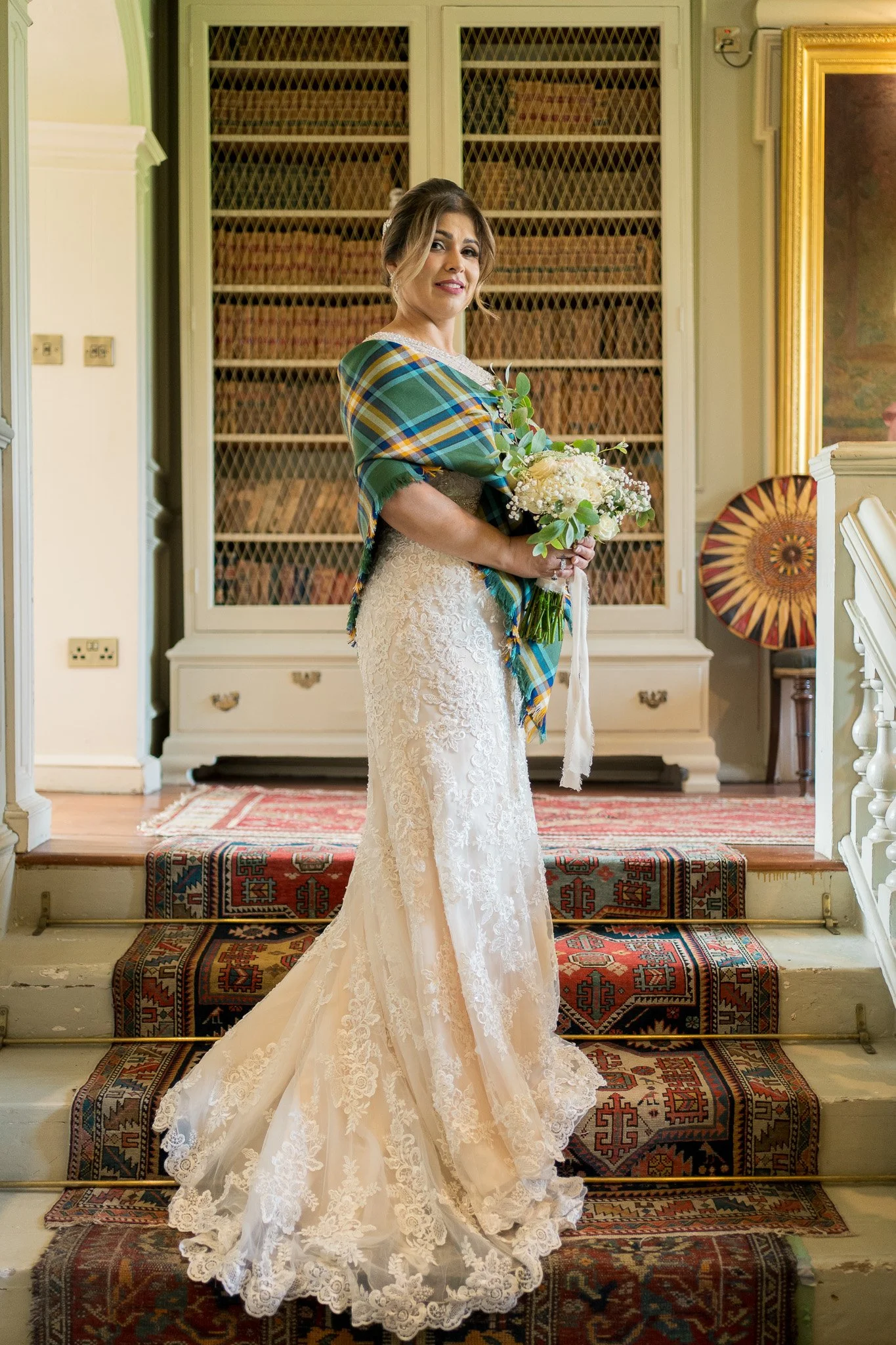 A woman in a white lace wedding dress holding a bouquet of white flowers, standing on a staircase with Persian rugs, in a vintage-style room with bookshelves and ornate furnishings.