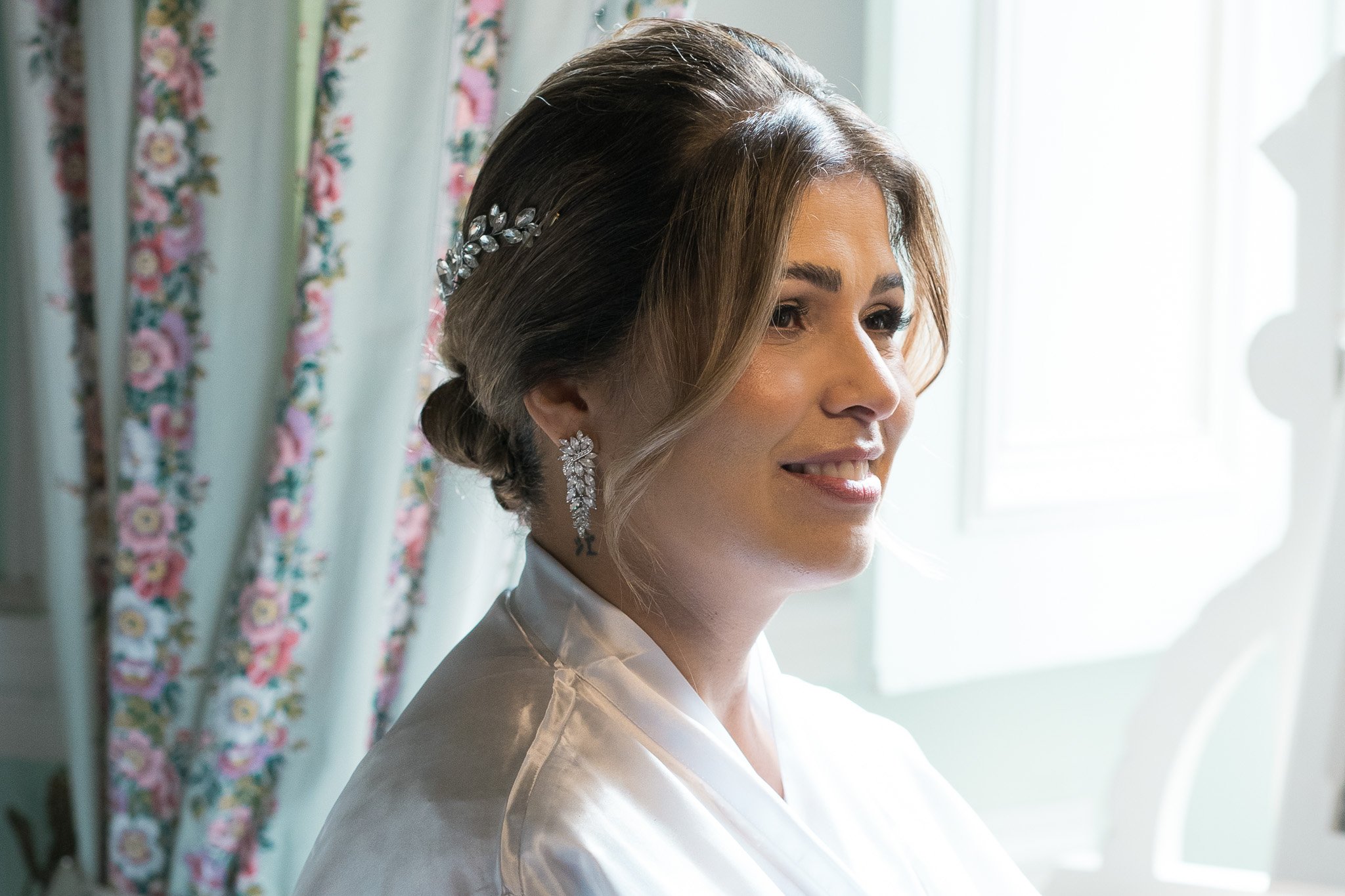 Bridal preparation with styled brown hair adorned with a decorative hair accessory, wearing elegant earrings, smiling gently while looking outside a window with floral curtain patterns in the background.