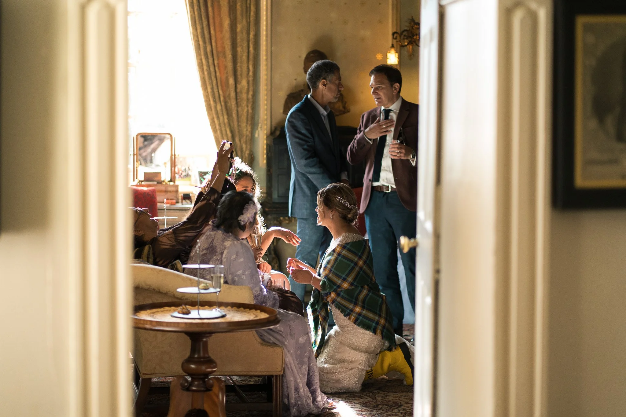 People having a celebration or gathering in a warmly lit room, some sitting on the floor in traditional clothing, while others stand and converse. The scene is viewed through a doorframe.