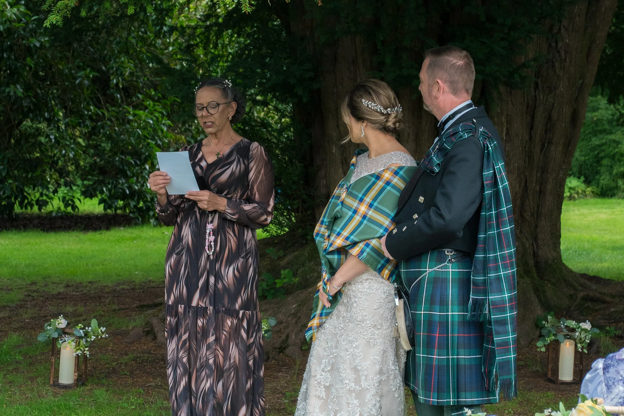 A wedding ceremony taking place outdoors under a large tree, with a woman reading from a paper, a bride in a lace dress, and a groom in a plaid kilt listening.