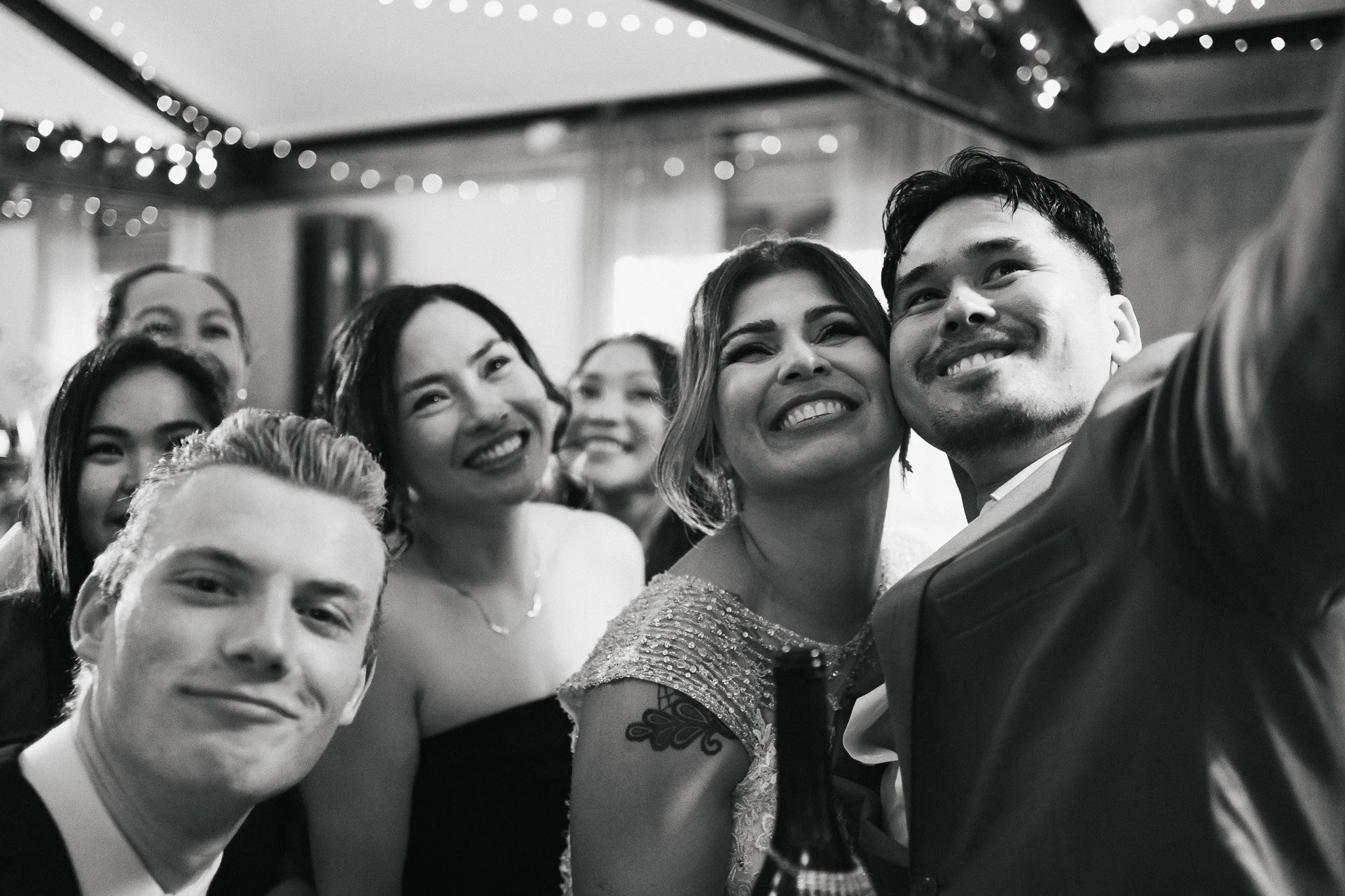 A group of diverse people smiling and taking a selfie at a celebration, with string lights in the background.