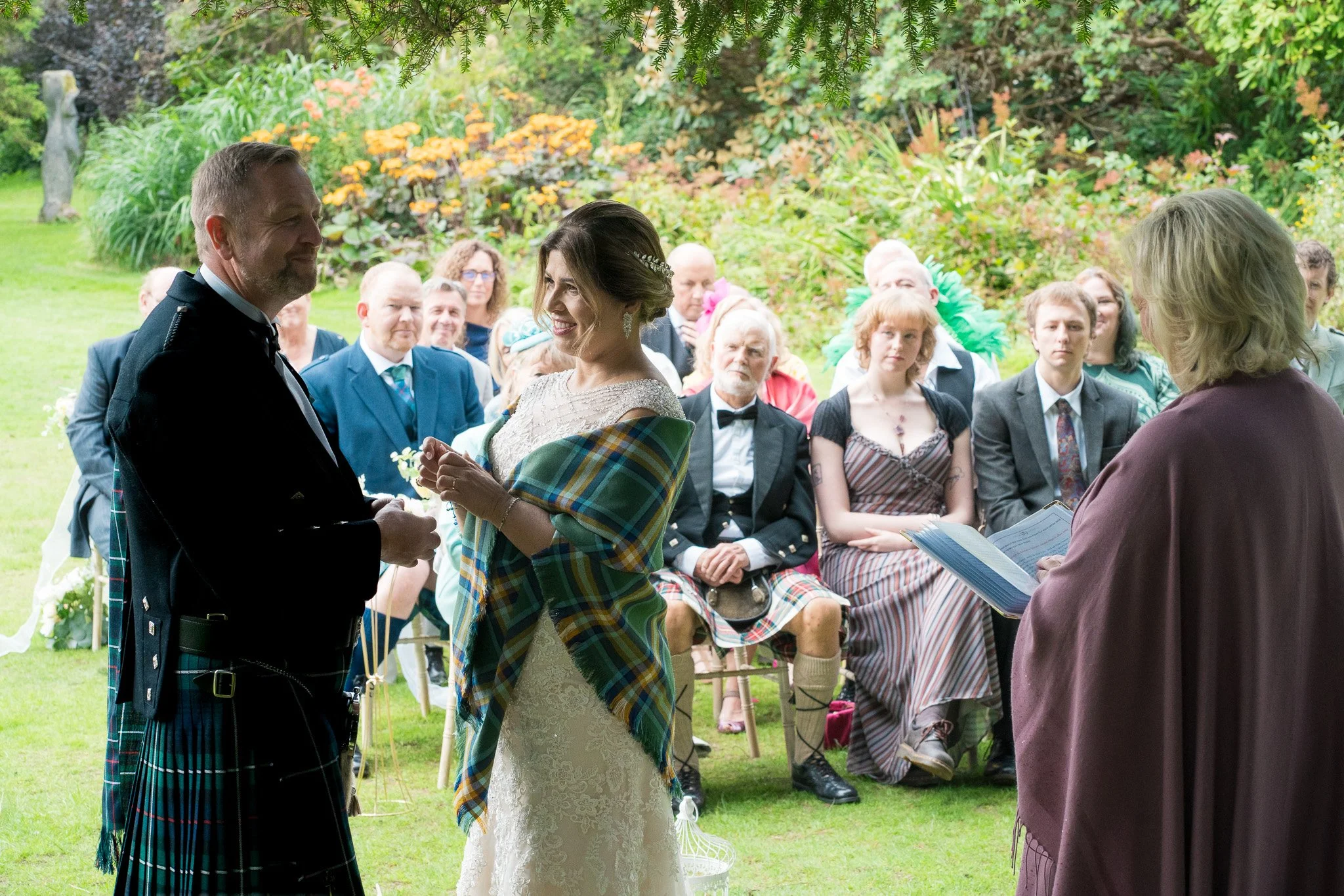 A bride and groom exchanging vows at a wedding ceremony outdoors, with guests seated and watching in a garden setting.