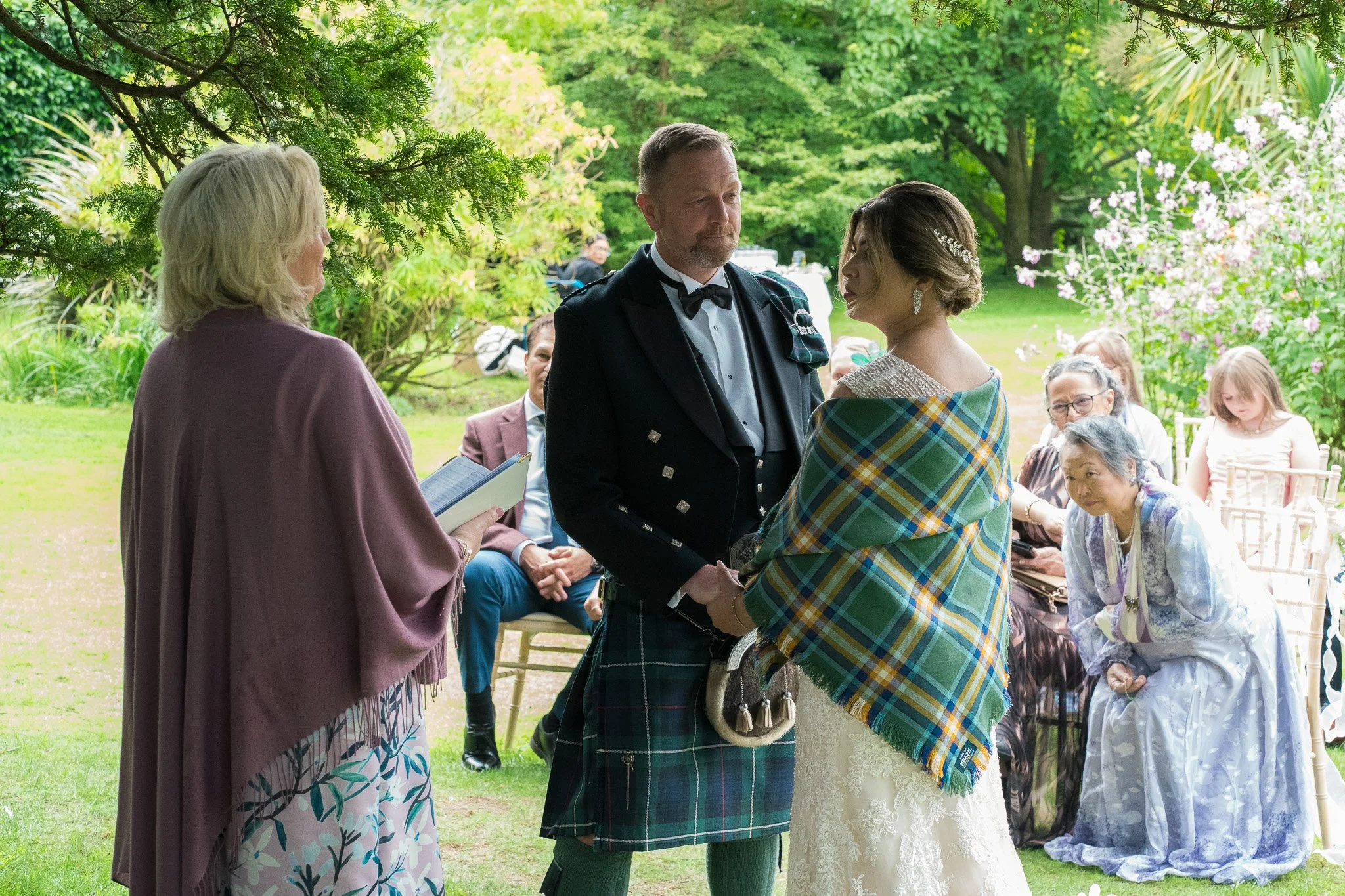 A wedding ceremony taking place outdoors in a lush garden. The bride and groom are exchanging vows, dressed in traditional attire, with the bride wrapped in a tartan shawl. A woman officiating the ceremony holds a book, and guests are seated behind t