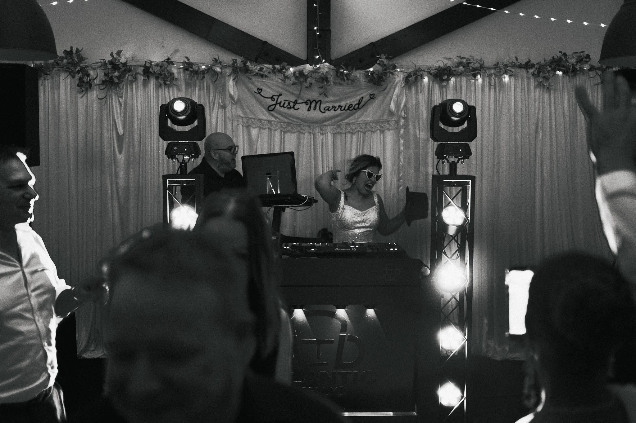 A woman celebrating at her wedding reception, wearing sunglasses and a dress, dancing with her arms raised, with a DJ playing music behind her. A banner that reads 'Just Married' is hanging in the background, decorated with flowers and hearts.