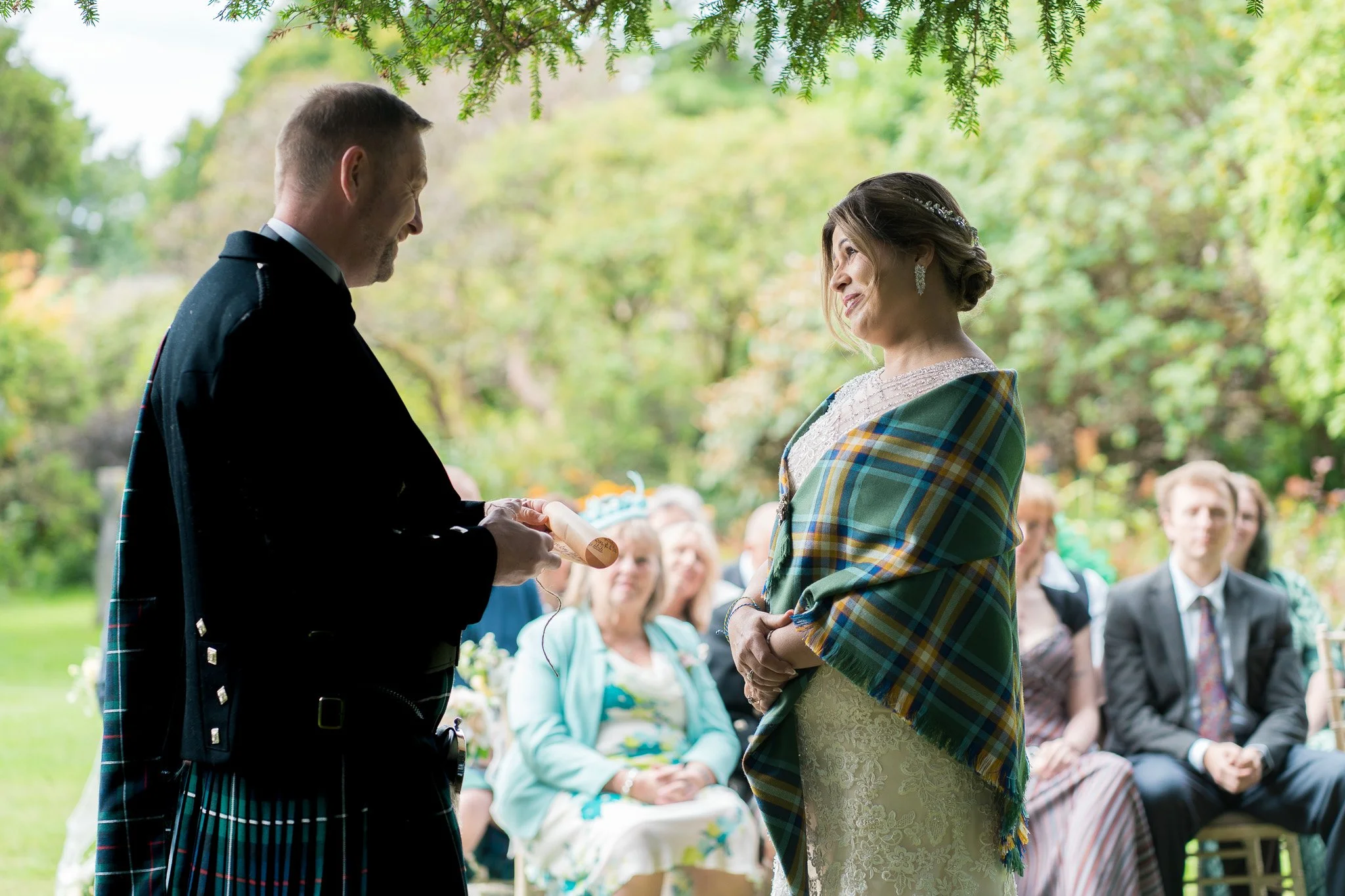 A man in a traditional Scottish kilt and a woman in a lace dress with a green plaid shawl exchange vows during an outdoor wedding ceremony, with guests seated and trees and greenery in the background.