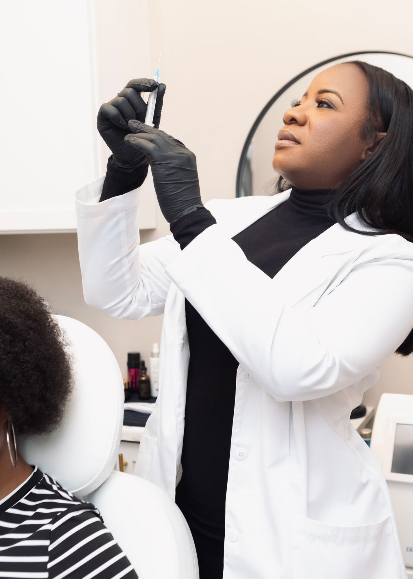 A healthcare professional in a white coat and black gloves preparing a syringe, with a patient sitting in a chair nearby in a medical office.