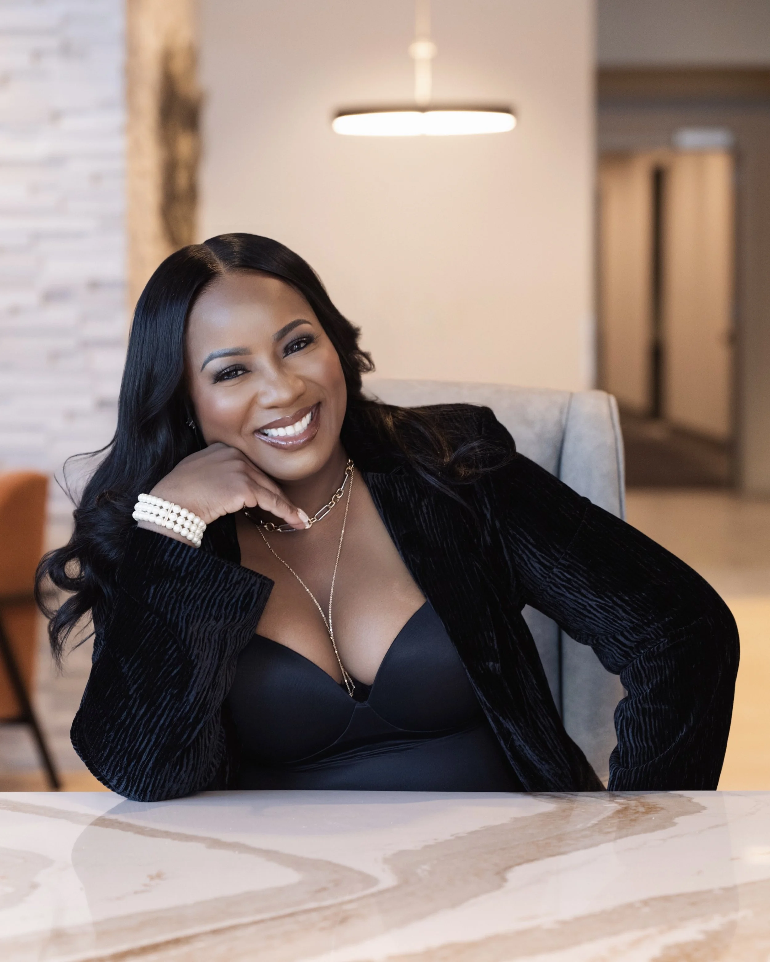 A woman with long black hair, wearing a black top and black velvet blazer, smiling and sitting at a marble table in a modern, well-lit room.