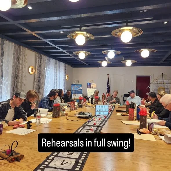 People sitting around a long conference table during a rehearsal, with a miniature train track running along the center of the table, and a sign that reads 'Week of Angels' on the train track.