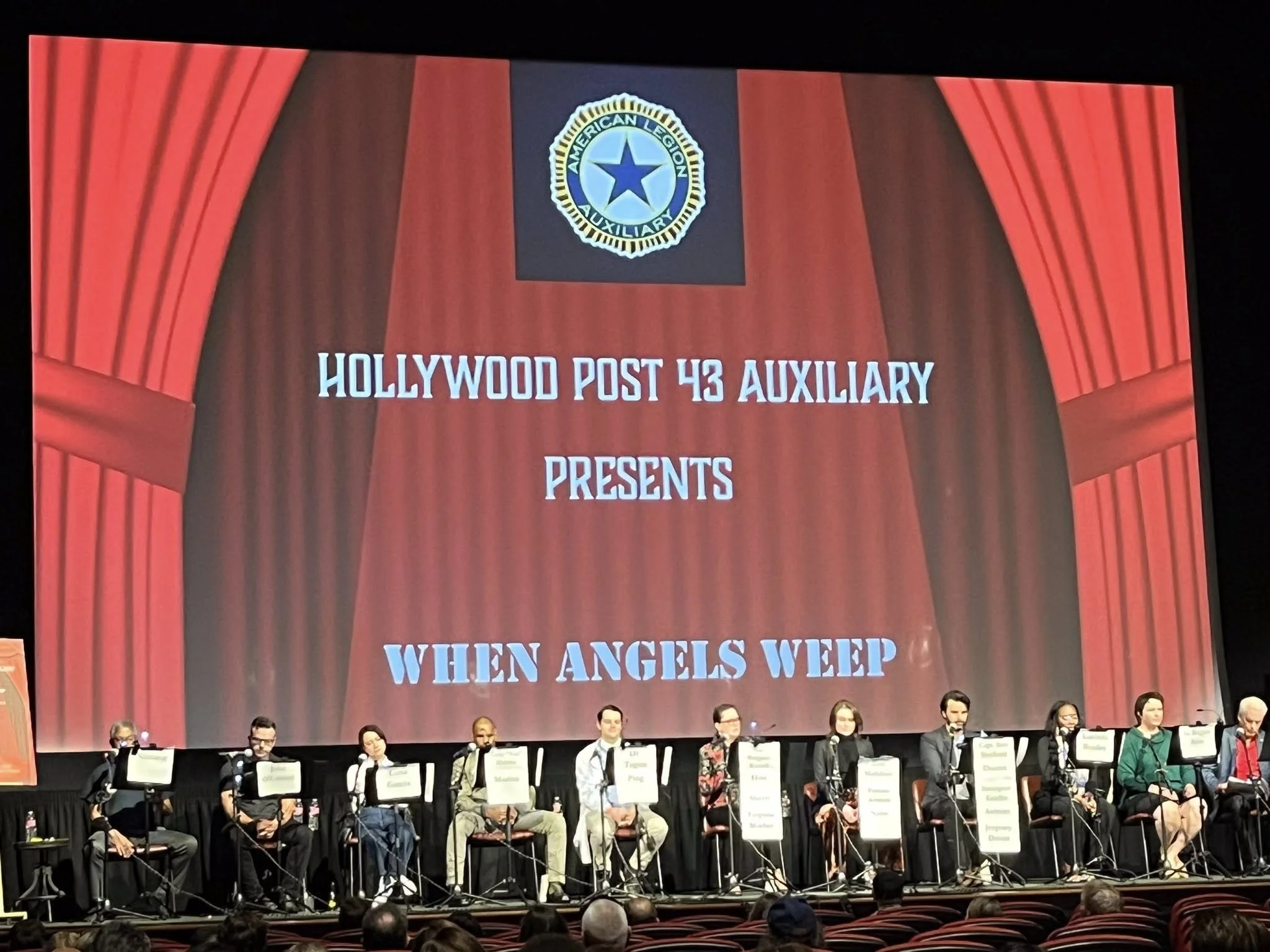 A stage with a large screen displaying red curtains with the American Legion Auxiliary emblem at the top, and text reading "Hollywood Post 43 Auxiliary Presents When Angels Weep." There are ten people seated at tables with microphones in front of them.