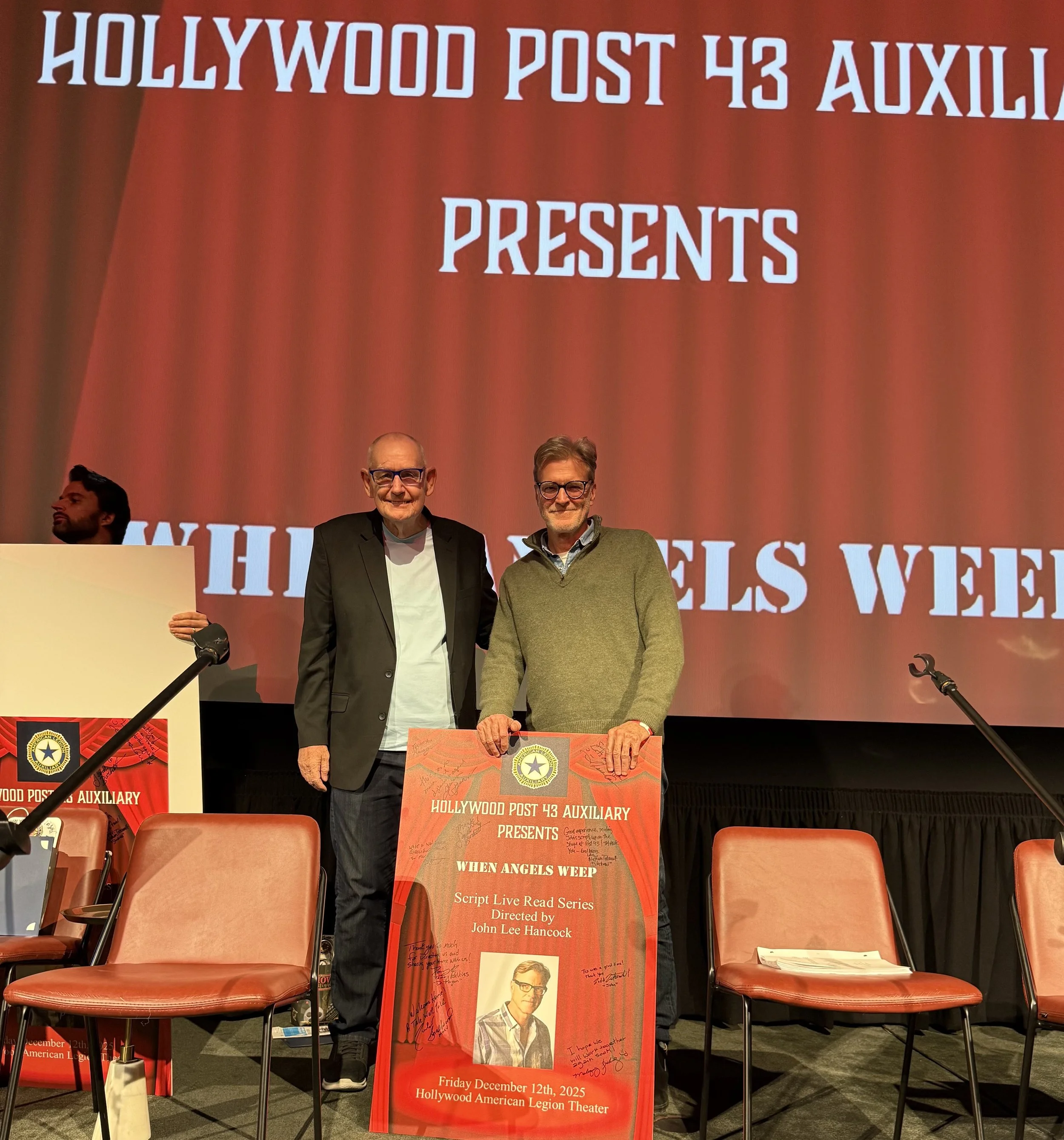 Two men stand on stage with a large red backdrop that reads 'Hollywood Post 43 Auxiliary Presents When Angels Weep.' One man wears a black blazer, glasses, and a white t-shirt, the other man wears a green sweater and glasses. They are holding a framed poster for a script read series titled 'When Angels Weep,' directed by John Lee Hancock, scheduled for December 12th, 2025, at the Hollywood American Legion Theater. Several empty chairs are visible on stage.