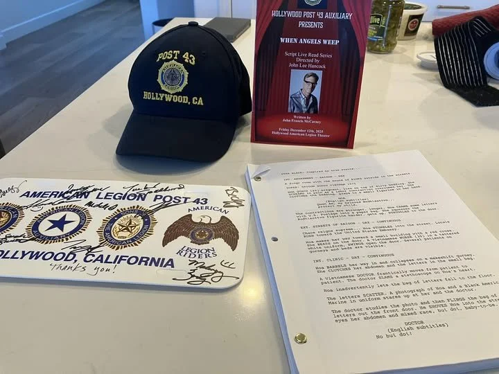 A navy blue baseball cap with yellow embroidery that reads "Post 43 Hollywood, CA". Next to it, a red pamphlet titled "When Angels Weep" with the Hollywood Post 43 Auxiliary presents. Also on the table, a white paper with signatures and handwritten notes, and some typed pages. In the background, there are green pills or containers and a striped black and red item.