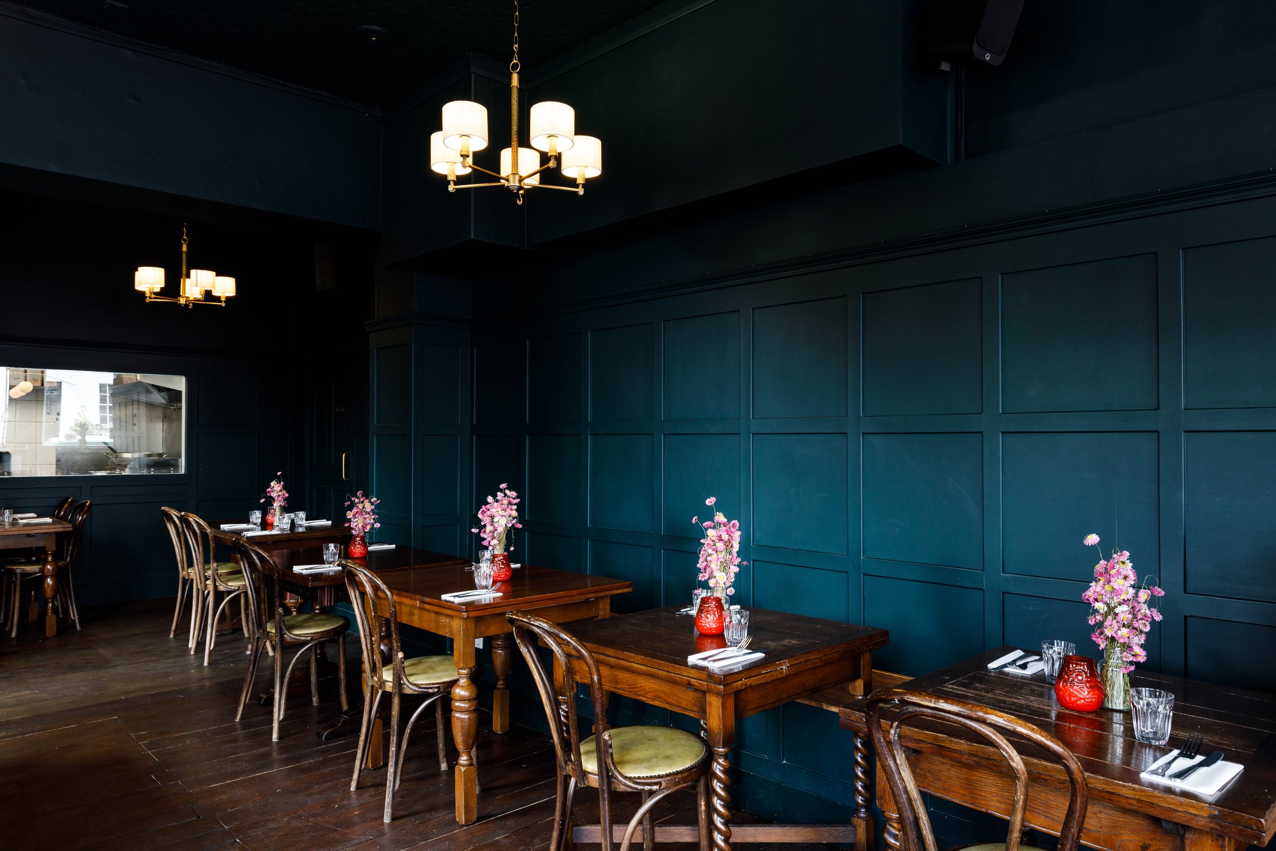 A dining room with dark blue walls and wooden tables set with glassware, napkins, and vases filled with pink flowers. There are wooden chairs around the tables and a large window on the left side. Two chandeliers with multiple light bulbs hang from the ceiling.