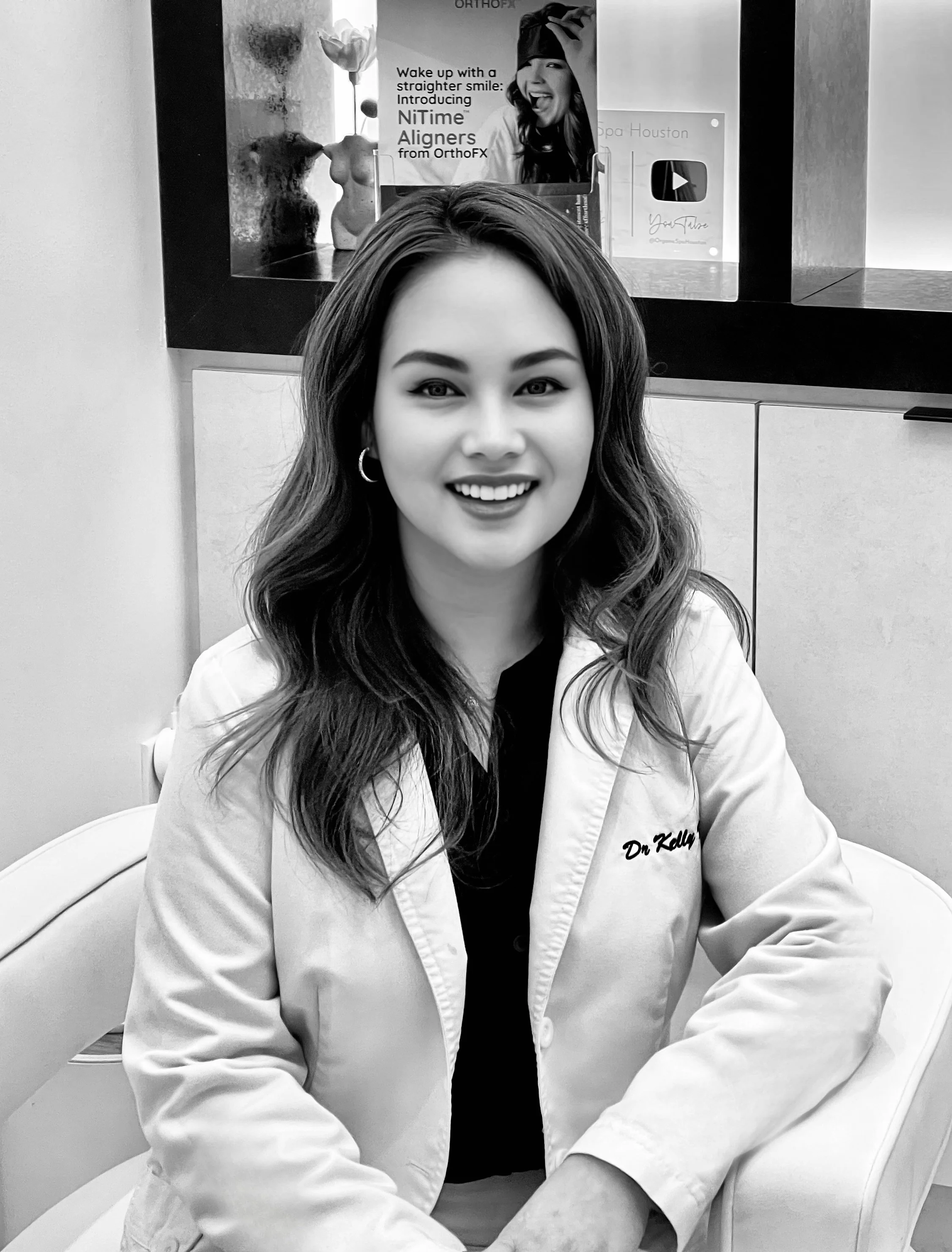 A young woman with long wavy hair and hoop earrings sitting in a white chair, smiling at the camera. She is wearing a white lab coat with the name "Dr. Kelly" embroidered on it, in a professional or clinical setting.