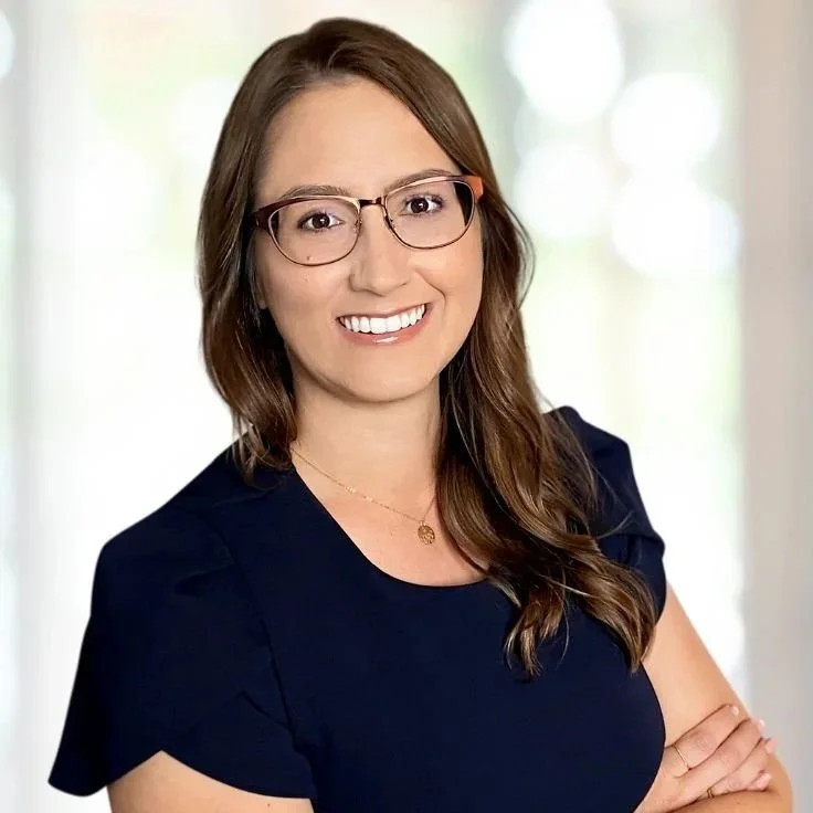 A young woman with long brown hair, wearing glasses and a navy blue top, smiling with crossed arms against a blurred indoor background.