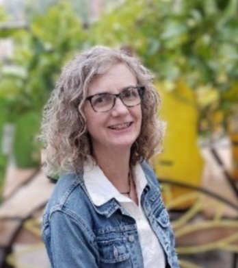 Woman with curly gray hair and glasses smiling outdoors, wearing a denim jacket and white shirt, with green foliage in the background.