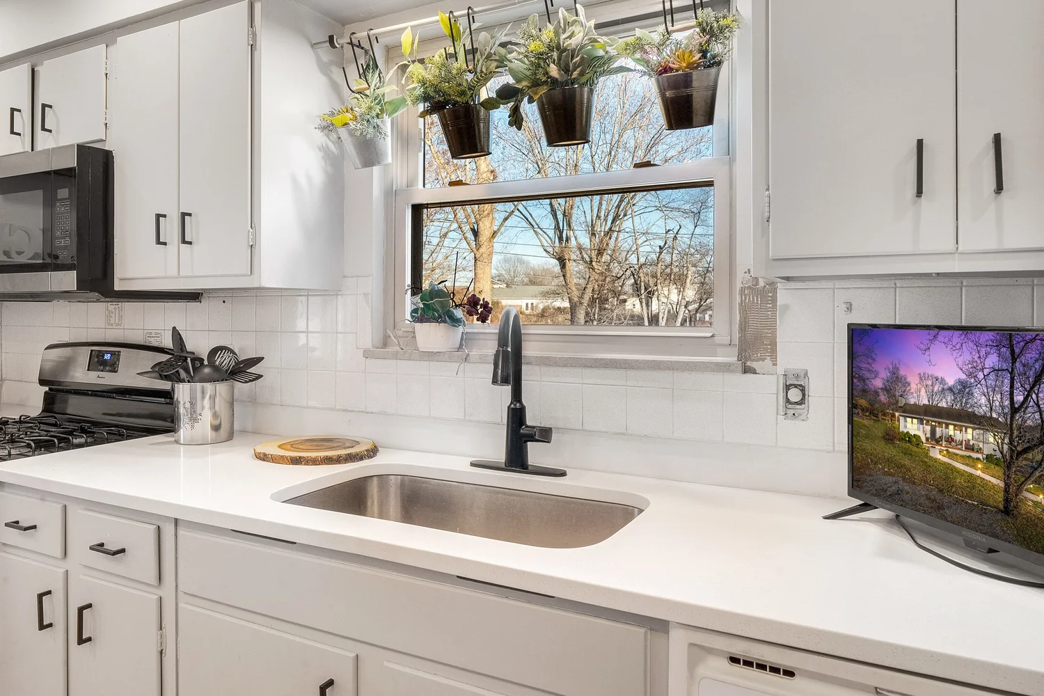 A white kitchen sink and dark faucet beneath a window with fresh flowers.