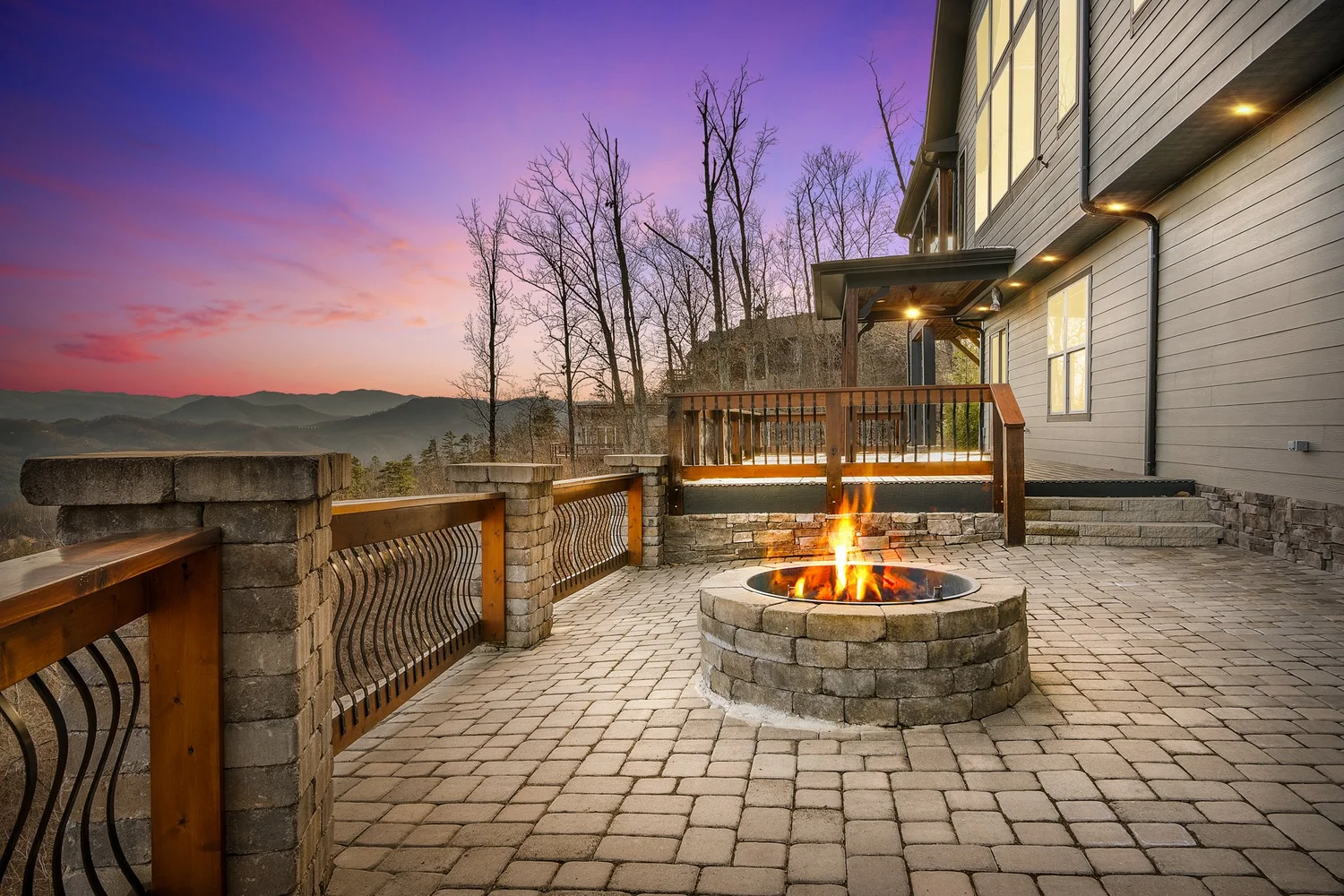 A stone terrace with a lit firepit beside a home and mountain views at twilight.