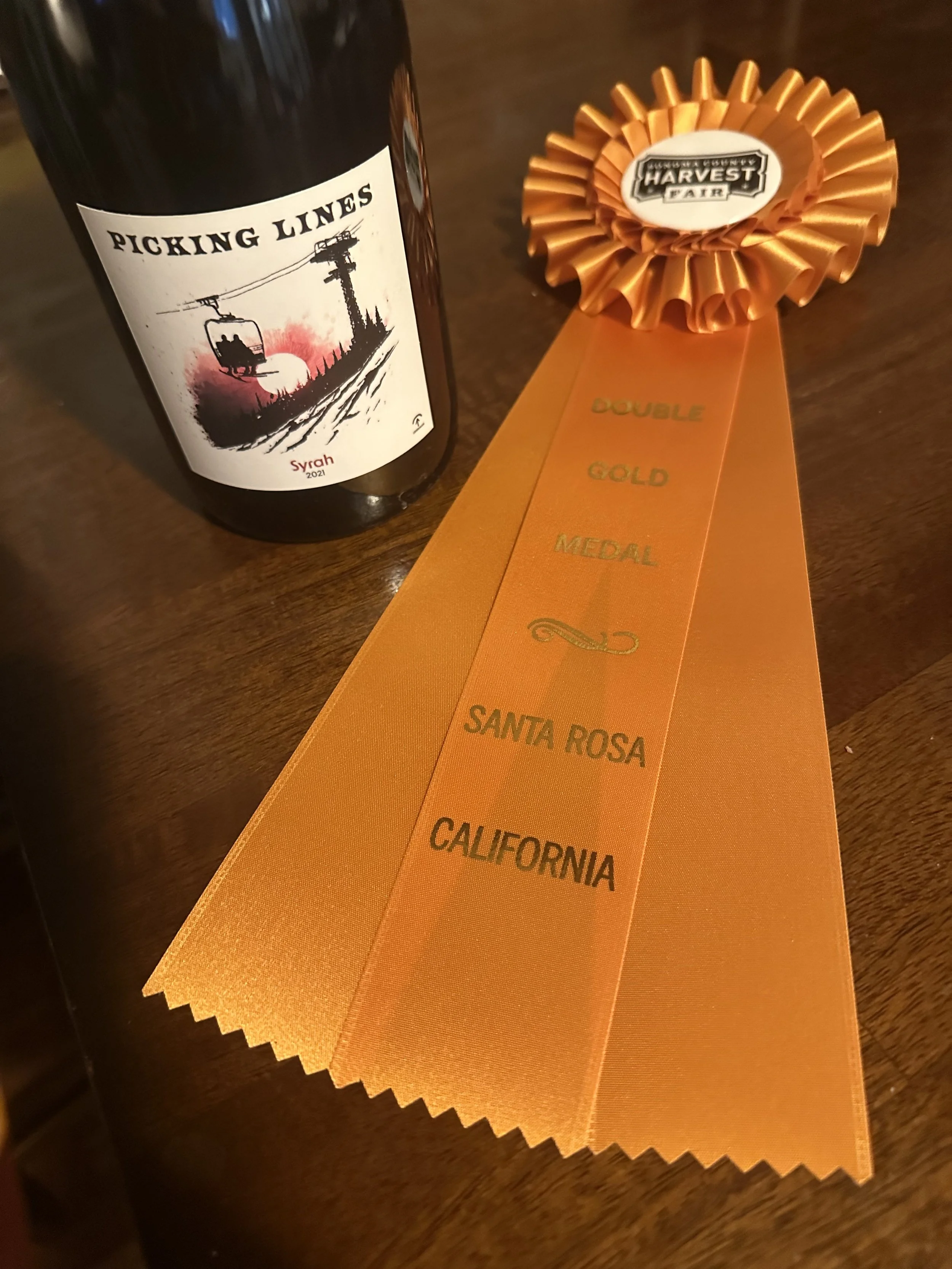 A bottle of Picking Lines Syrah wine and an orange award ribbon with a rosette that has a Harvest Fair logo, and the ribbon reads 'Double Gold Medal Santa Rosa California'.