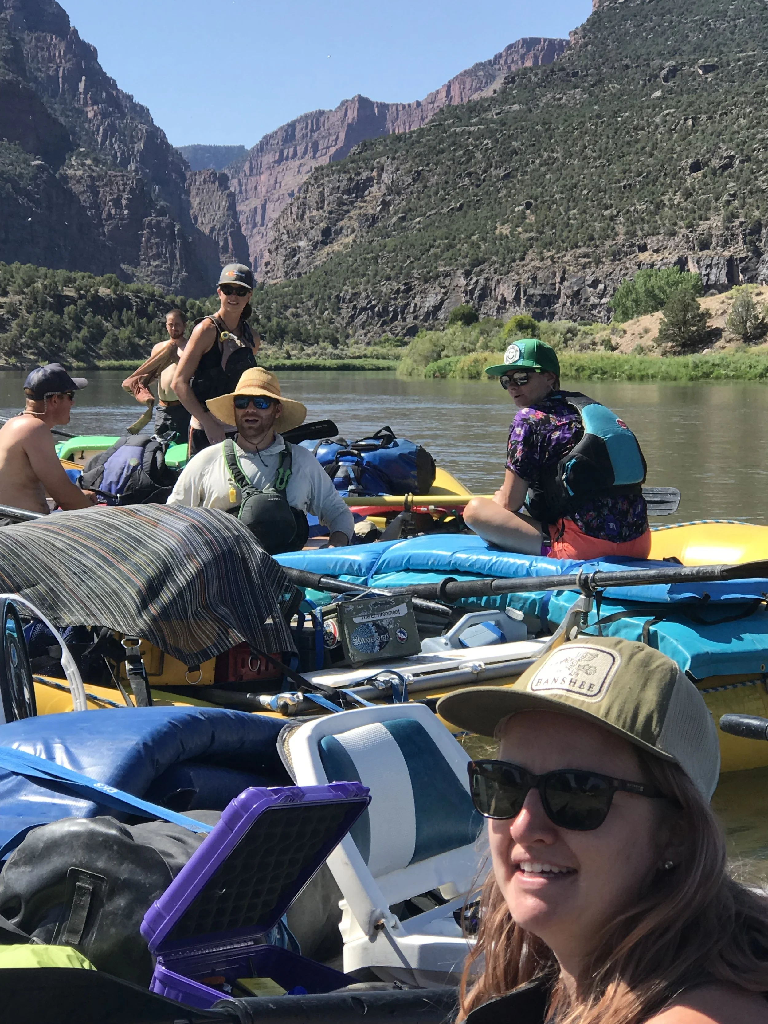 A group of people with kayaks and gear on a river under a mountainous landscape during daytime.