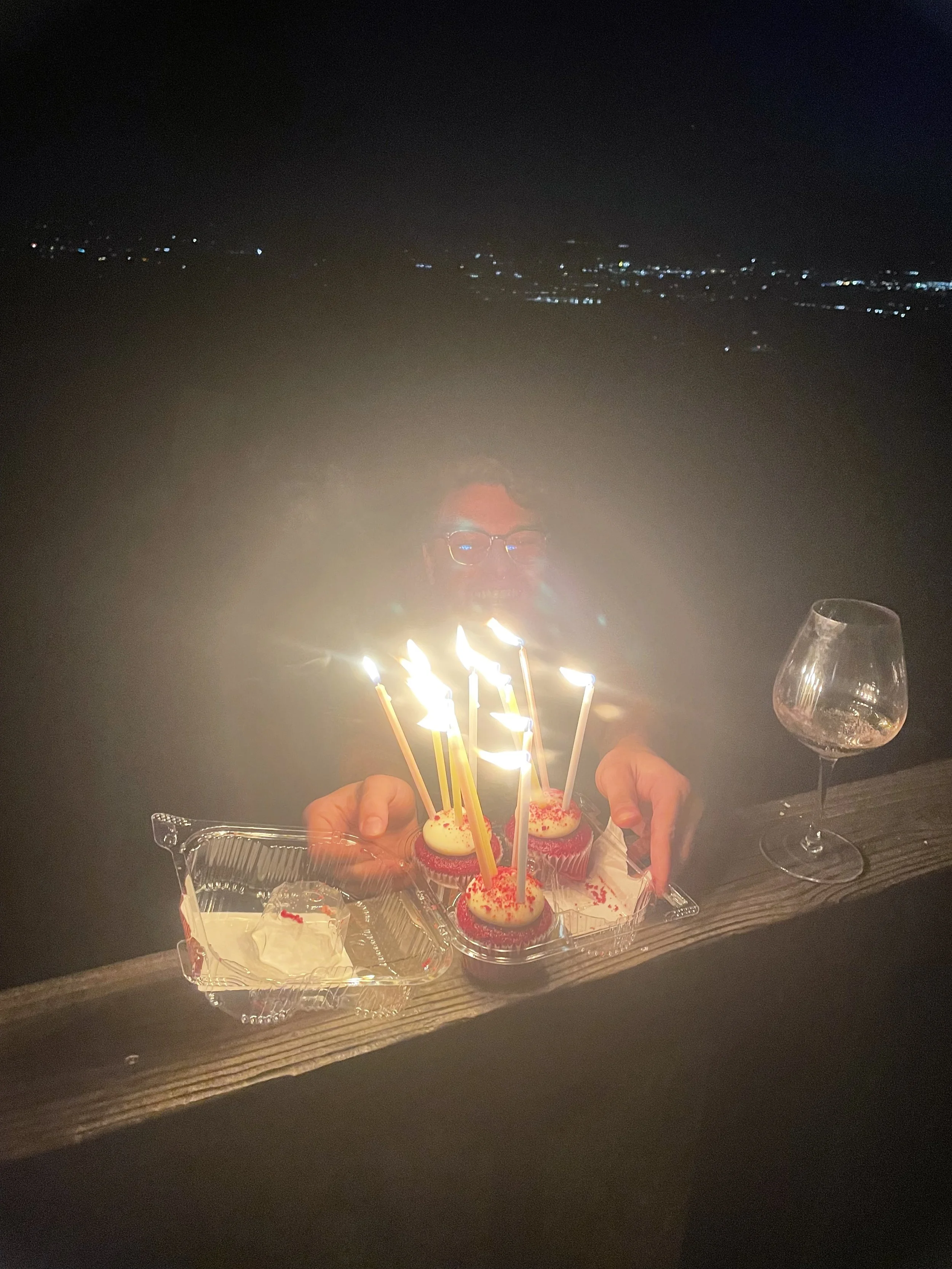 Person with glasses celebrating birthday with cupcakes and birthday candles at night, outdoors, with city lights in the background.