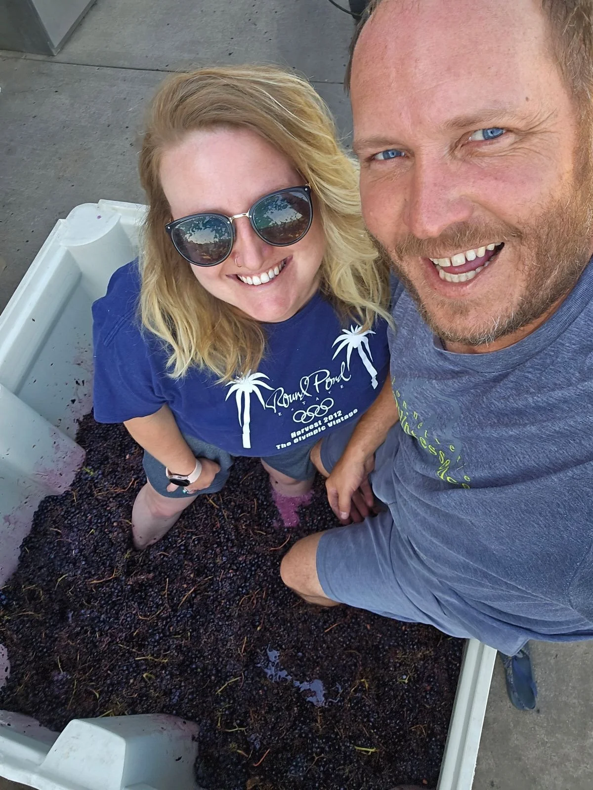 A smiling man and woman taking a selfie, standing in a large container filled with dark berries. The woman has blonde hair, is wearing sunglasses and a blue T-shirt, and the man has light-colored hair, a beard, and is wearing a gray T-shirt.