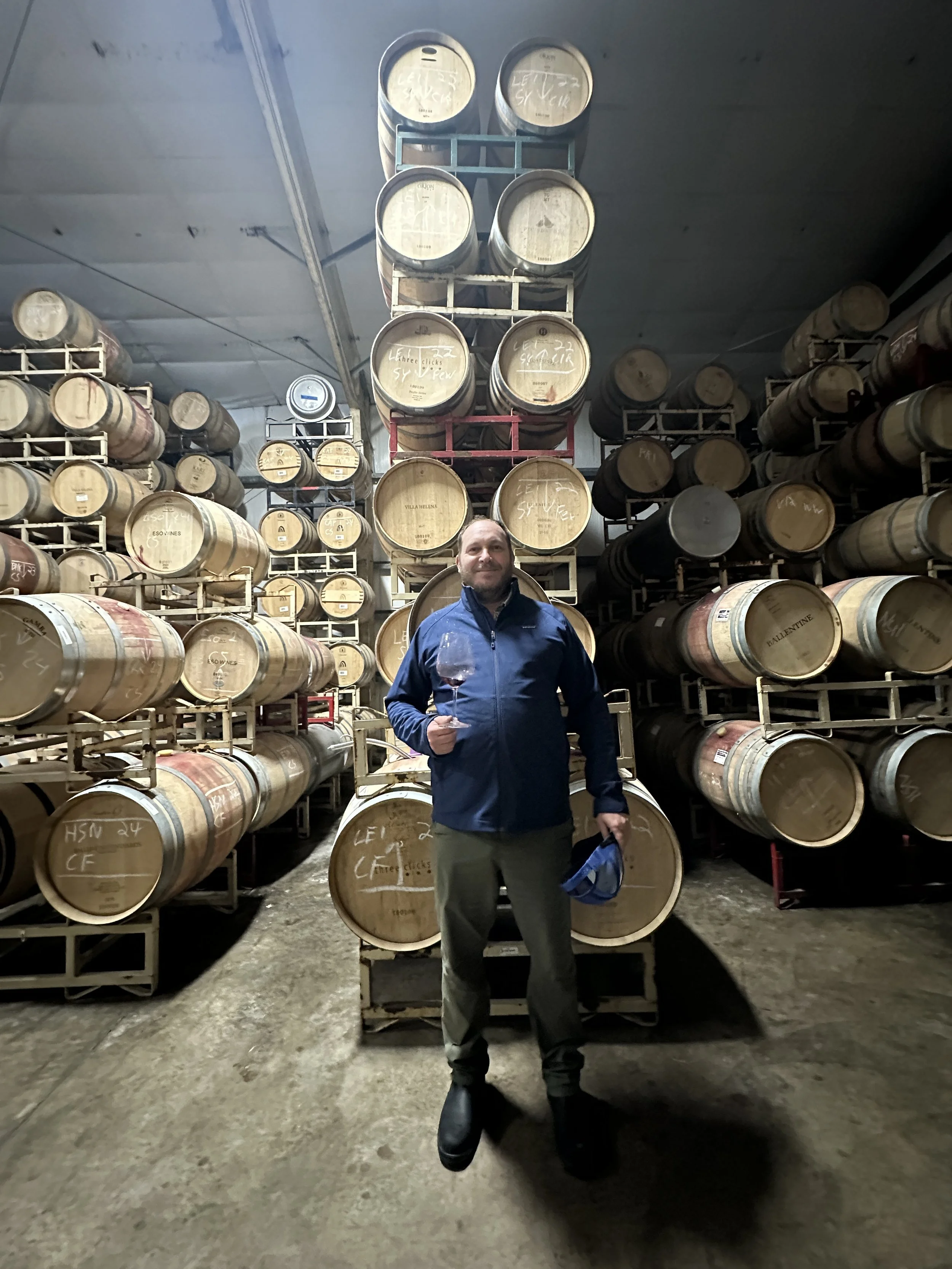 A man standing in a wine cellar with stacked wine barrels around him, holding a glass of red wine.