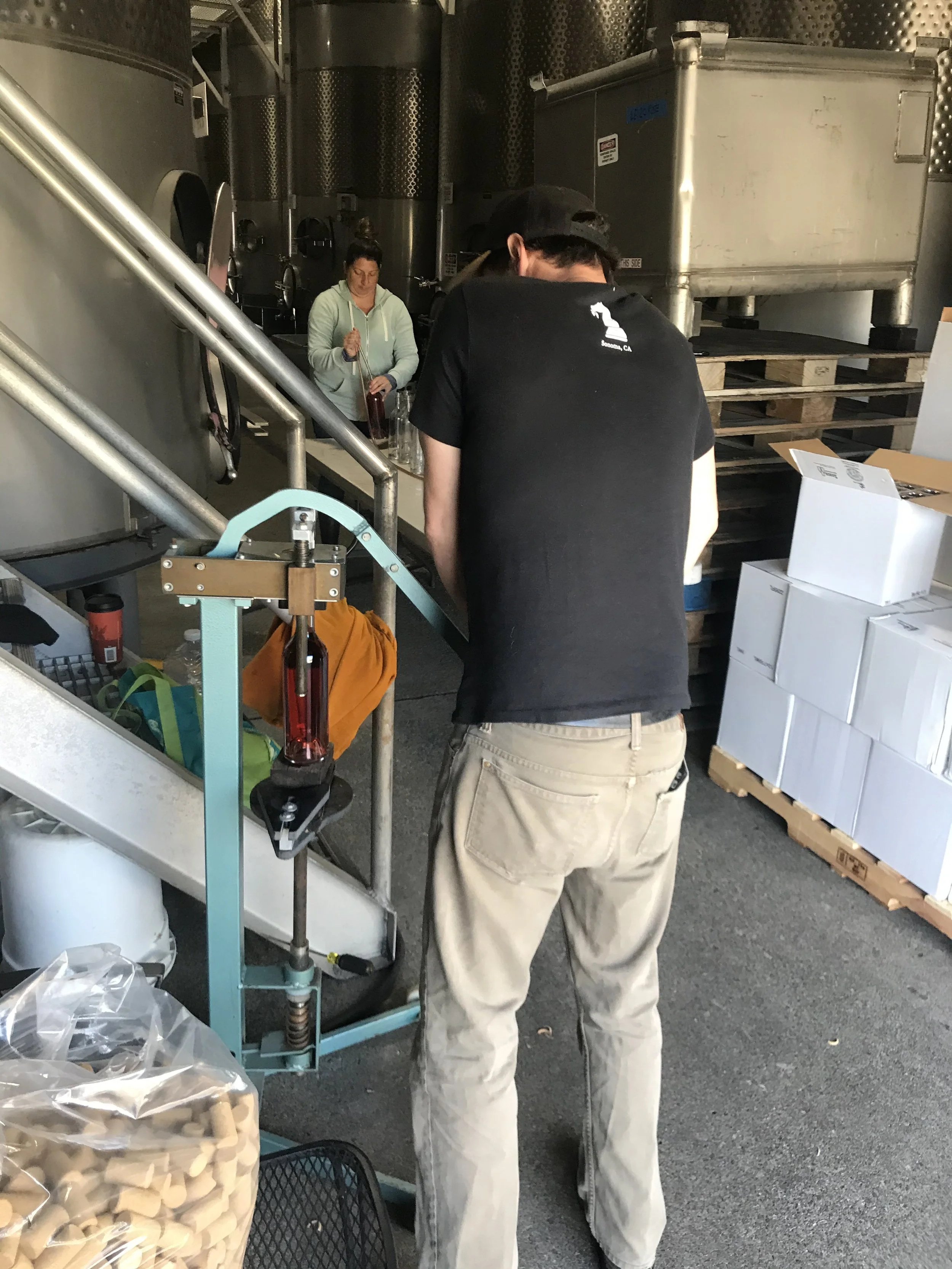 A man in a black t-shirt and beige pants working in a winery with large metal fermentation tanks in the background. A woman is seen in the background near the tanks.