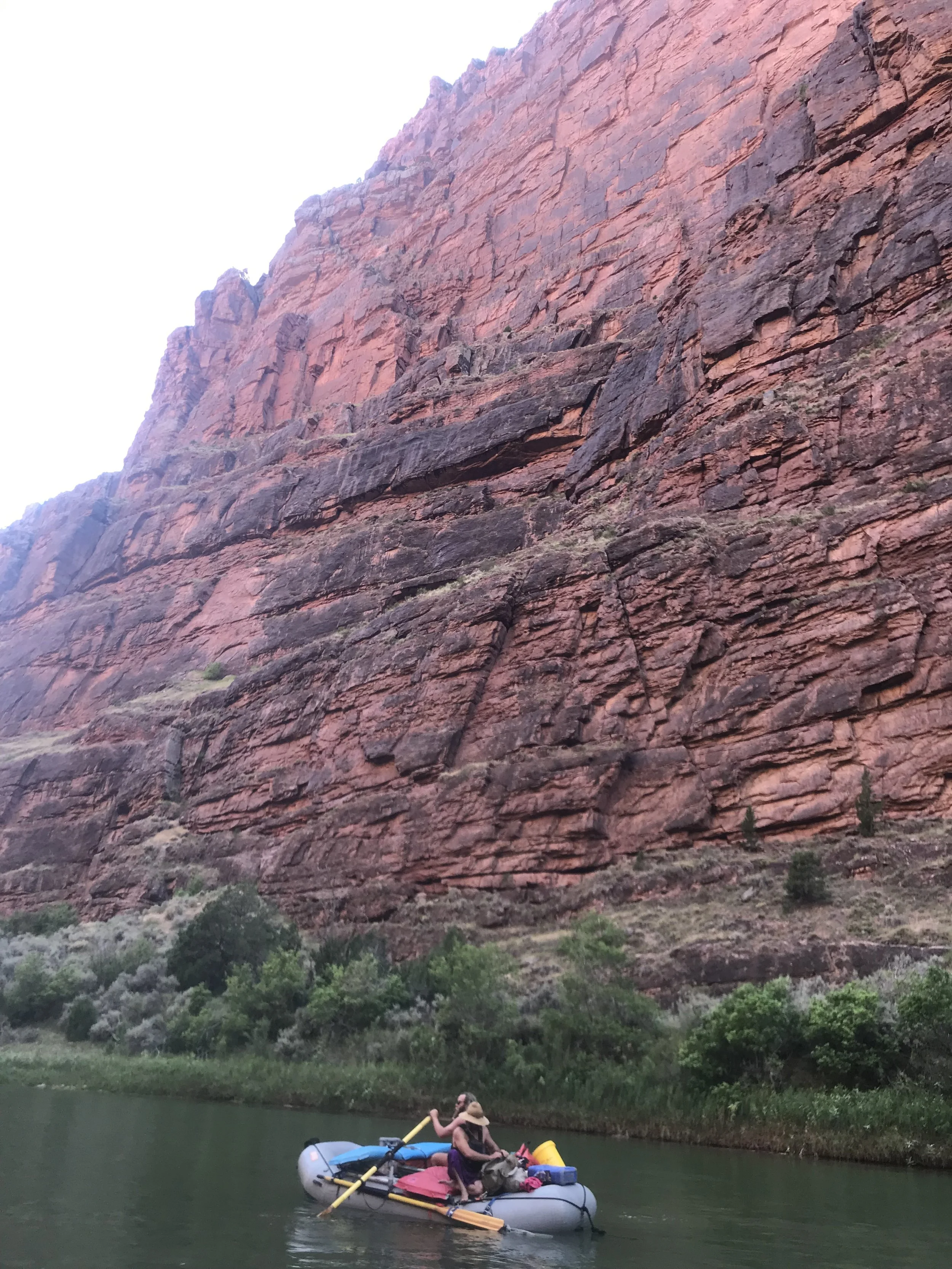People rafting on a river with a large red rock canyon wall in the background.