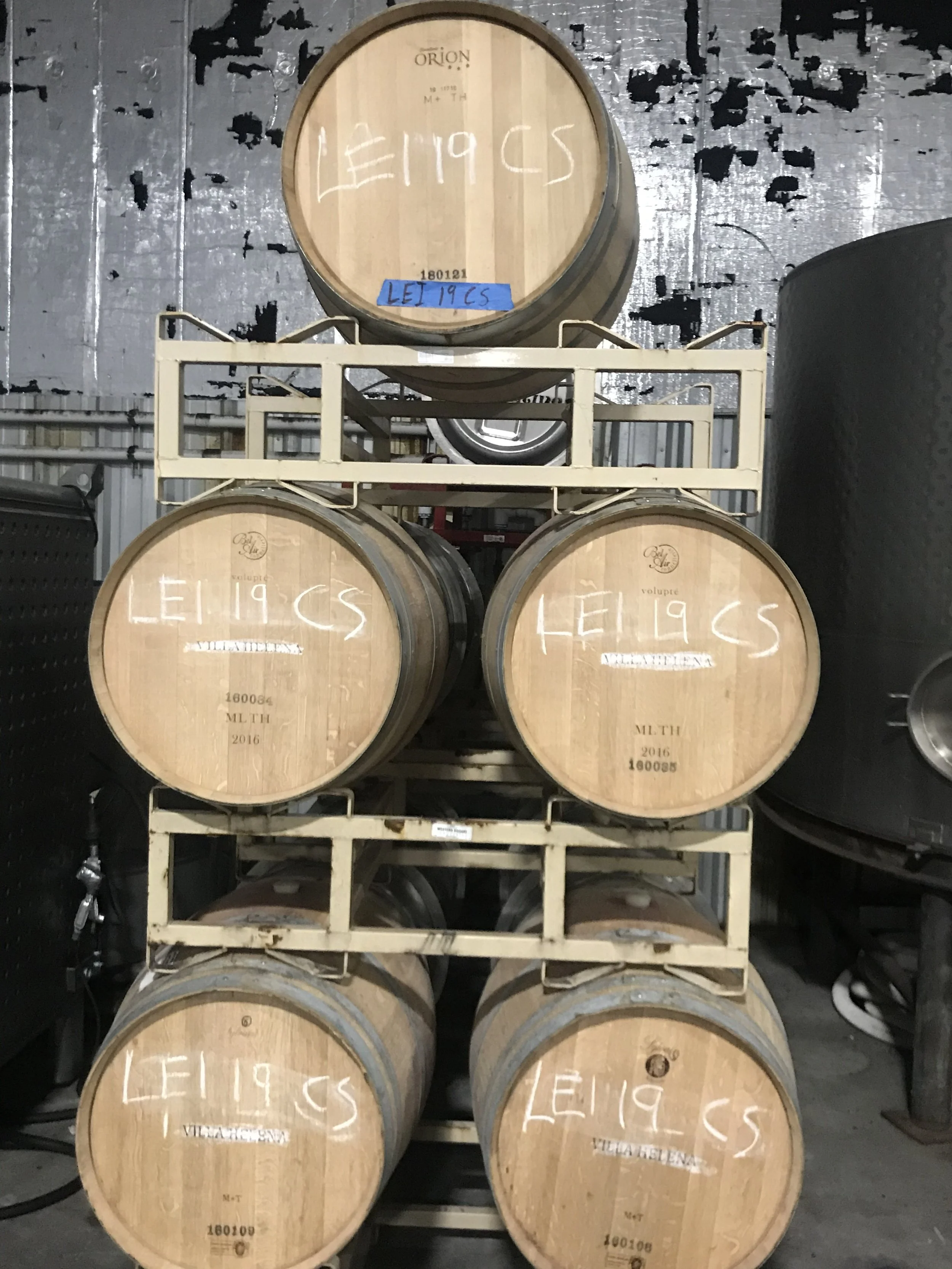 Six wooden wine barrels stacked in a storage rack, with labels reading 'LEI 19 CS' and other marking details, in a cellar or storage area.