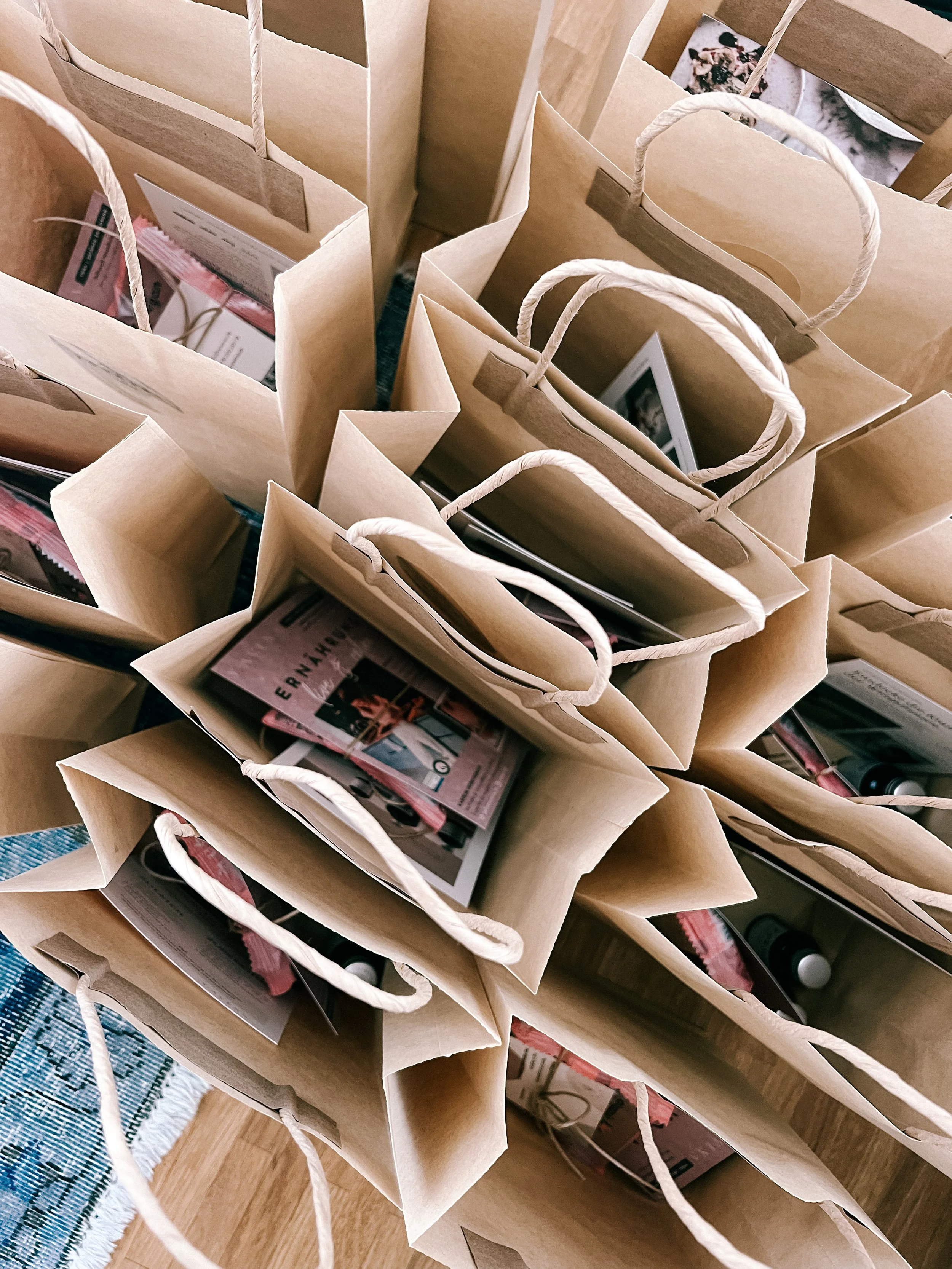 Mehrere braune Einkaufstüten mit weißen Griffen stehen auf einem Holzboden, einige Tüten enthalten Zeitschriften und andere Gegenstände.