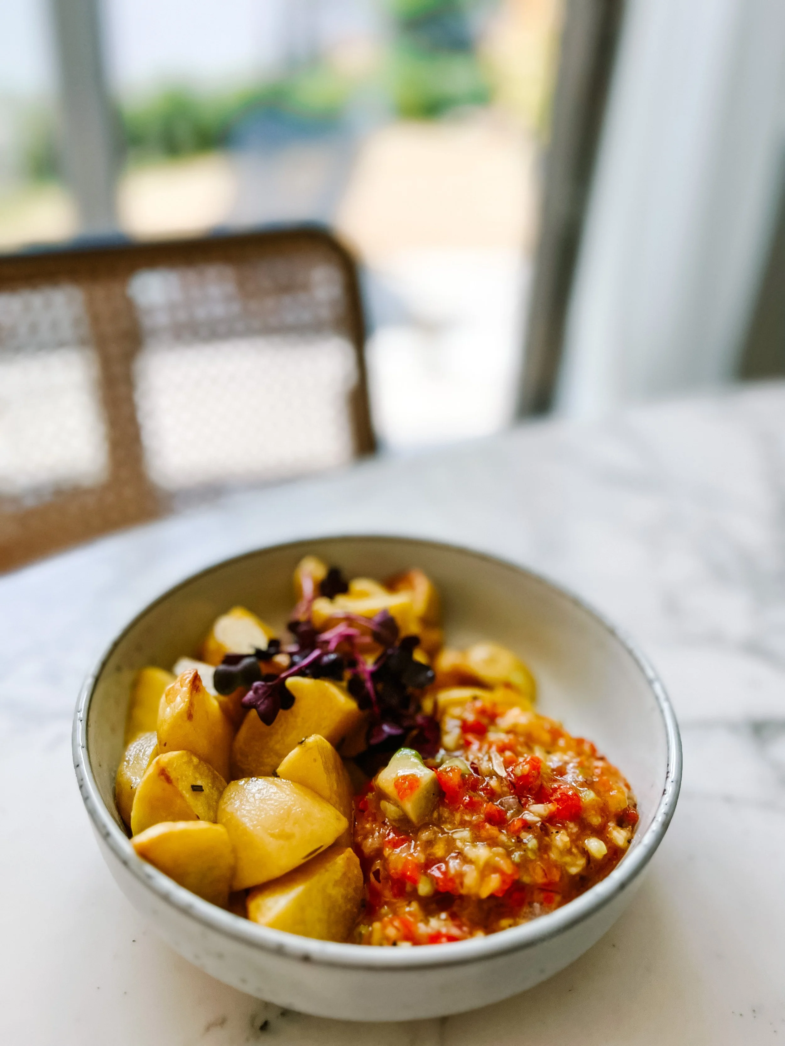 Schüssel mit gekocht gewürfelten Kartoffeln, einen Dip mit Tomaten und Zwiebeln, und eine Garnitur aus violetten Mikrogrün, auf einem weißen Tisch vor einem Fenster