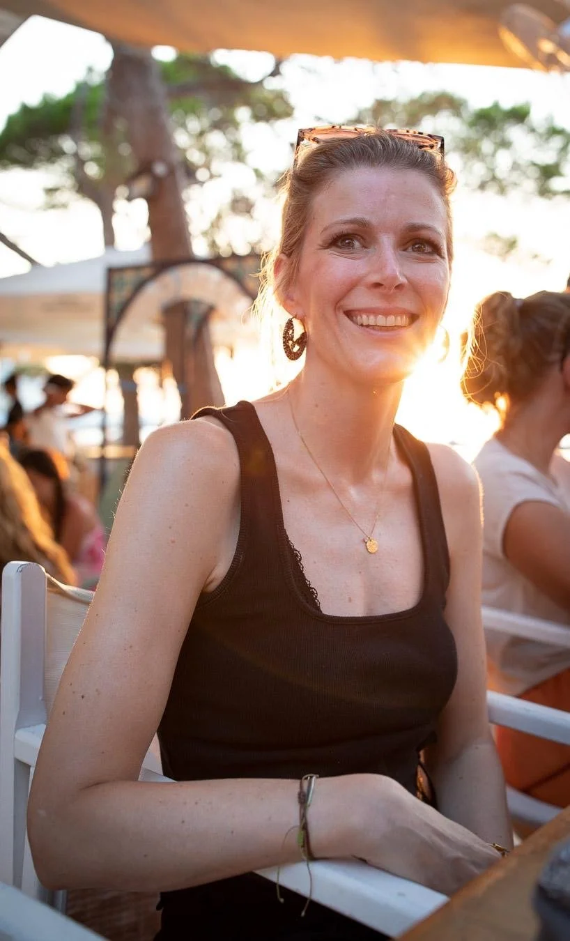 Eine lächelnde Frau mit braunen Haaren, Sonnenbrille auf dem Kopf, trägt ein schwarzes Top mit Spitze an den Schultern, sitzt an einem Tisch im Freien bei Sonnenuntergang, im Hintergrund sind andere Menschen und eine große Palme sichtbar.