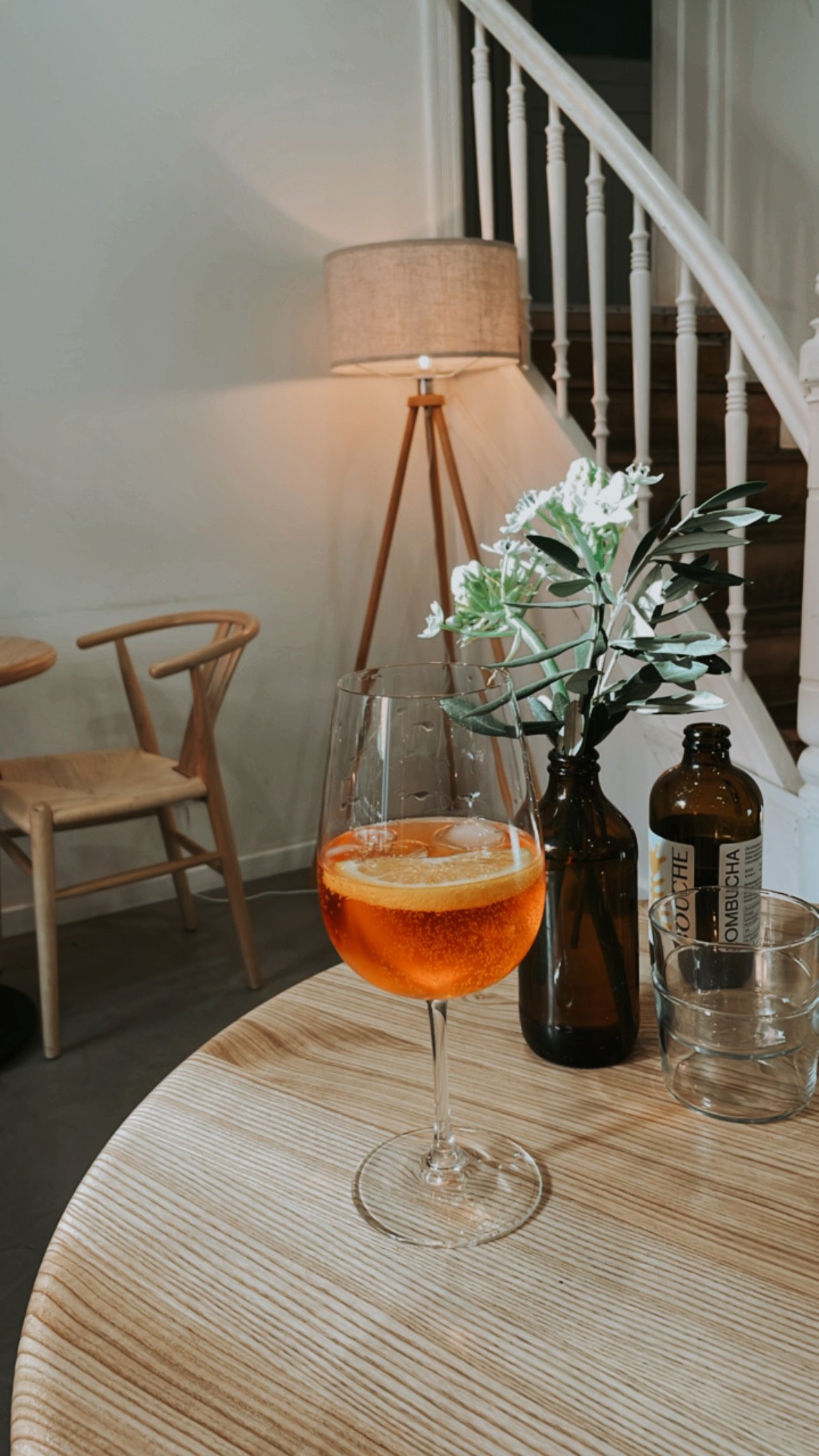 Ein Glase Aperol Spritz auf einem Holztisch mit Dekorationsflaschen und einer Vase mit weißen Blumen, im Hintergrund eine Stehlampe und eine Treppe.