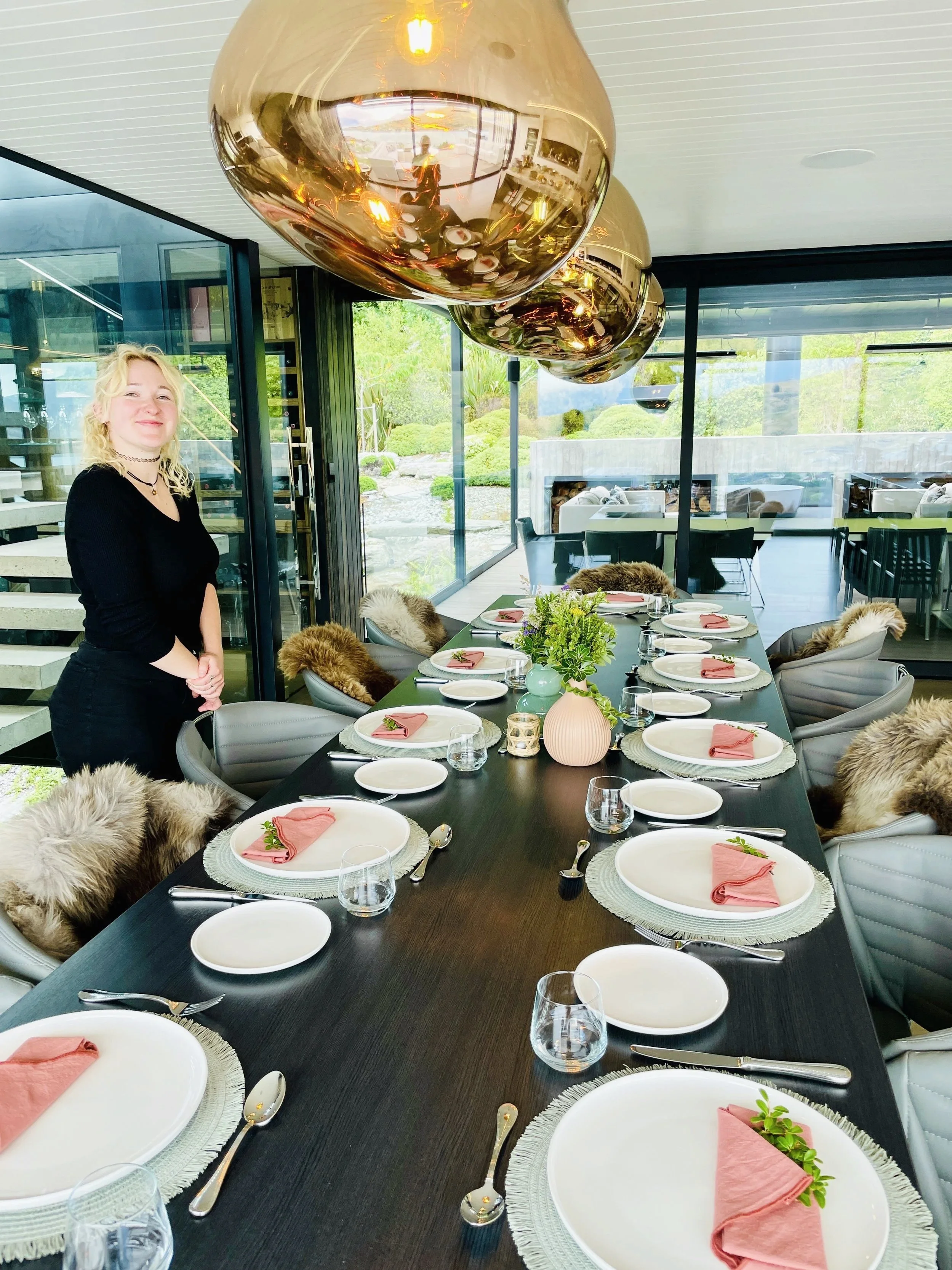 Queenstown Private Chef’s table styling -  a long dining table inside a modern room with large windows overlooking a garden. The table is set with white plates, rose coloured napkins, silverware, glasses, and a centerpiece with greenery and pink vase